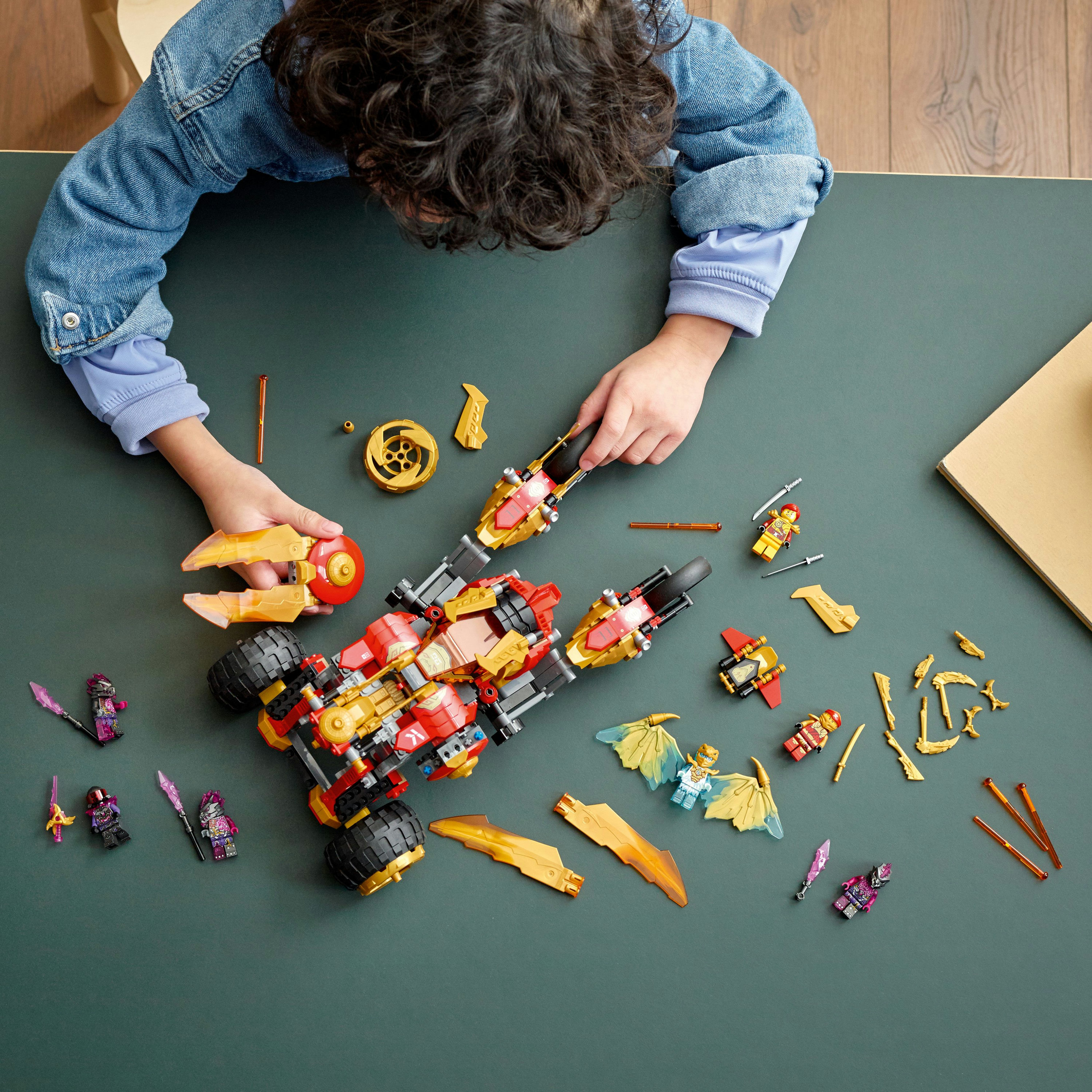 Un bambino assembla un veicolo giocattolo rosso e oro con varie figure e pezzi Lego su un tavolo verde.