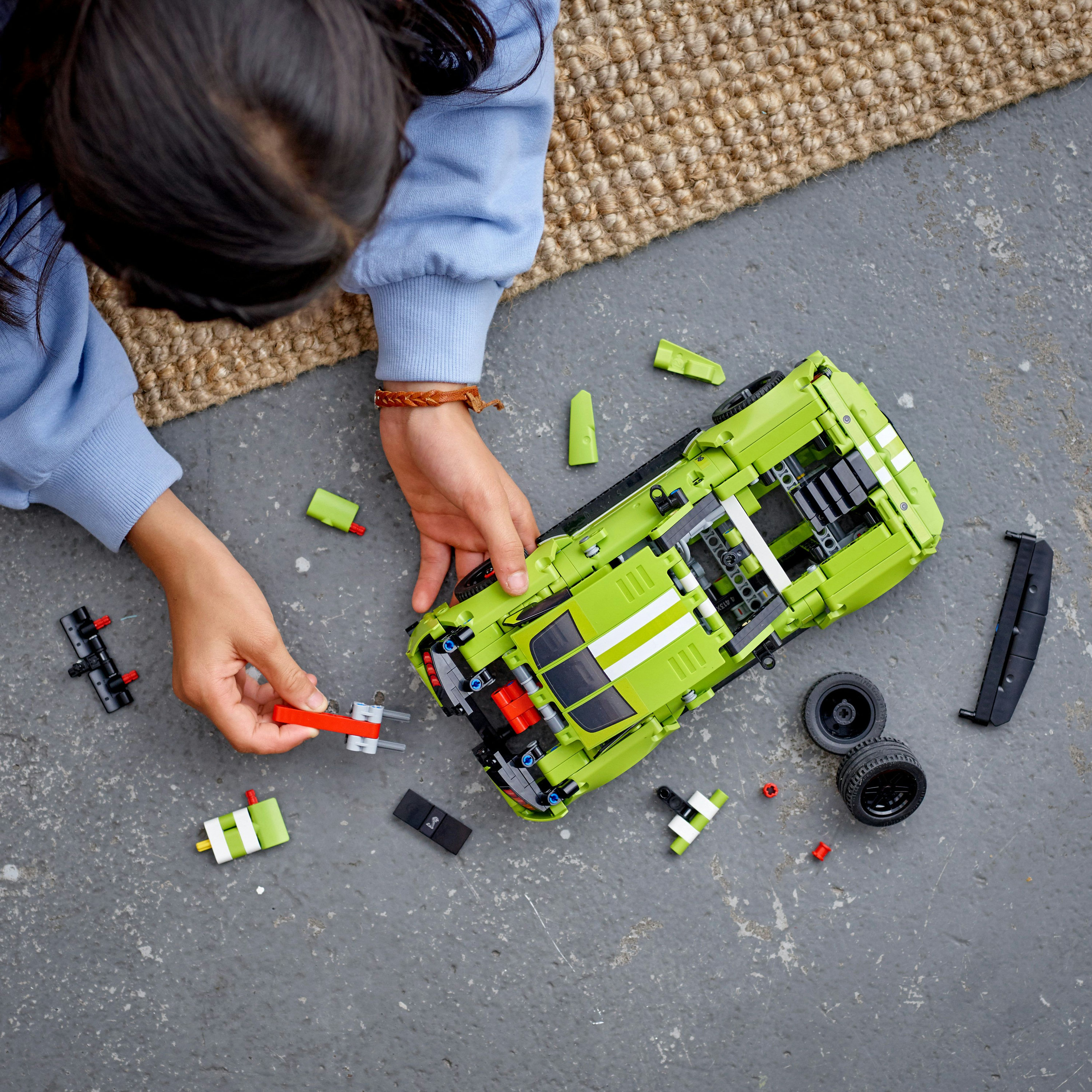 Ein Kind baut ein grünes Spielzeugauto zusammen. Teile des Autos und andere Teile liegen auf der grauen Oberfläche verstreut.