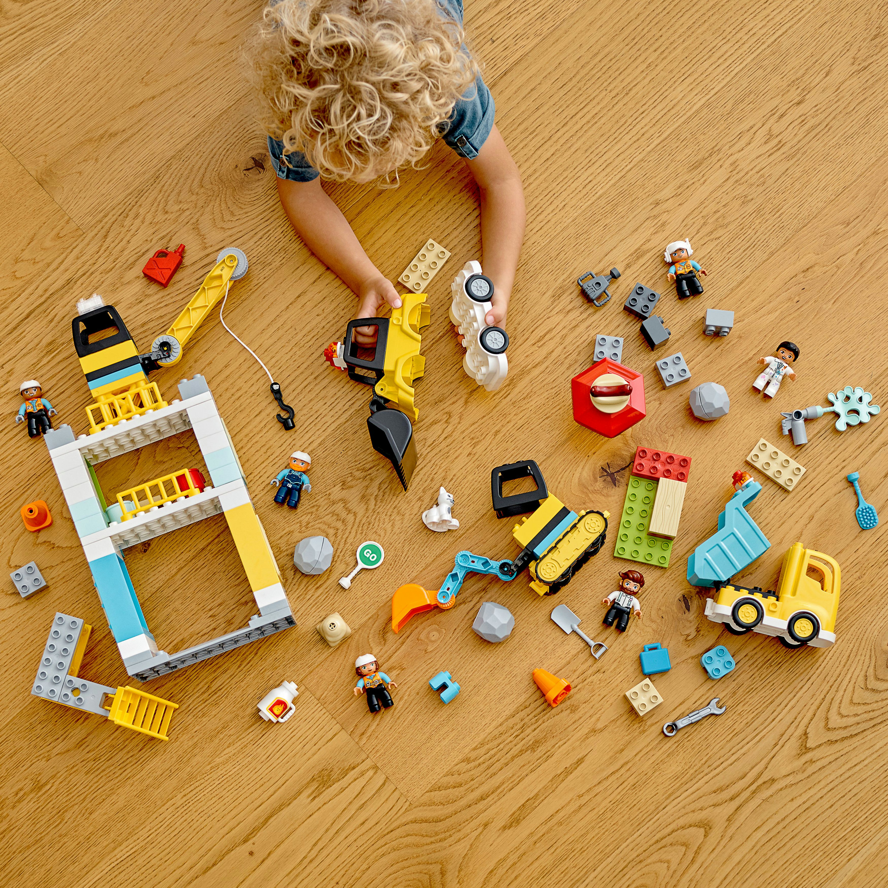 Dziecko bawi się zabawkowymi pojazdami budowlanymi i figurkami na drewnianej podłodze. Rozrzucone elementy.