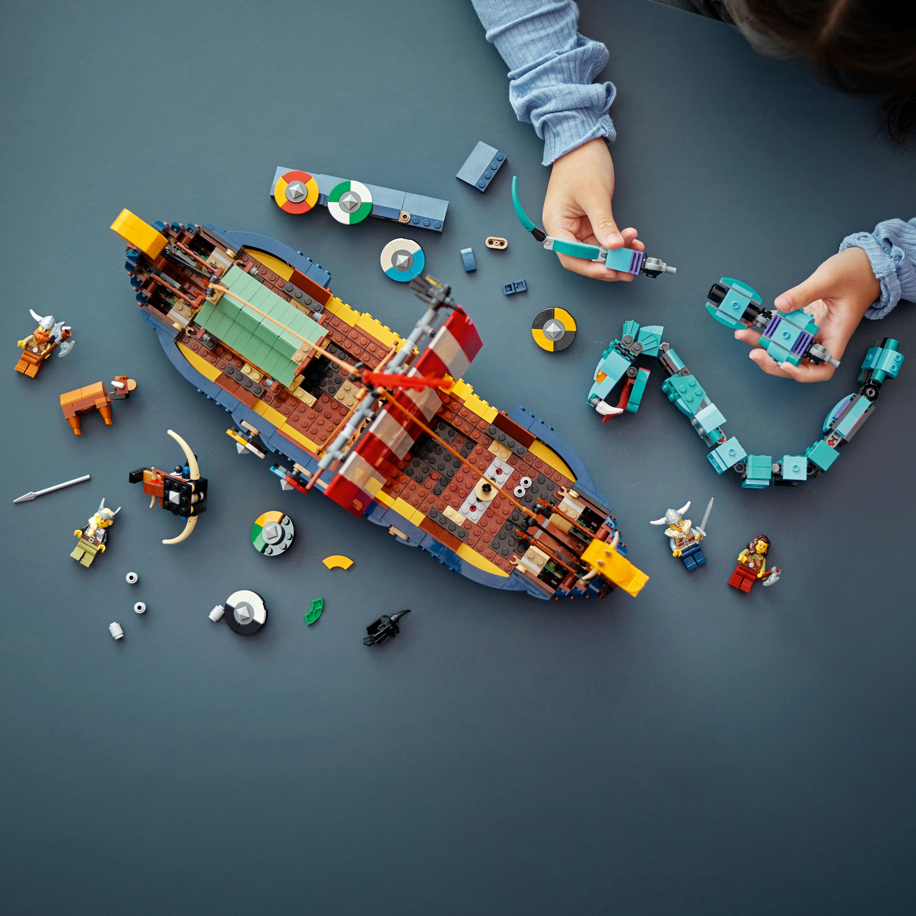 Handen bouwen een Lego-schip met minifiguren en diverse onderdelen op een donkerblauwe ondergrond.