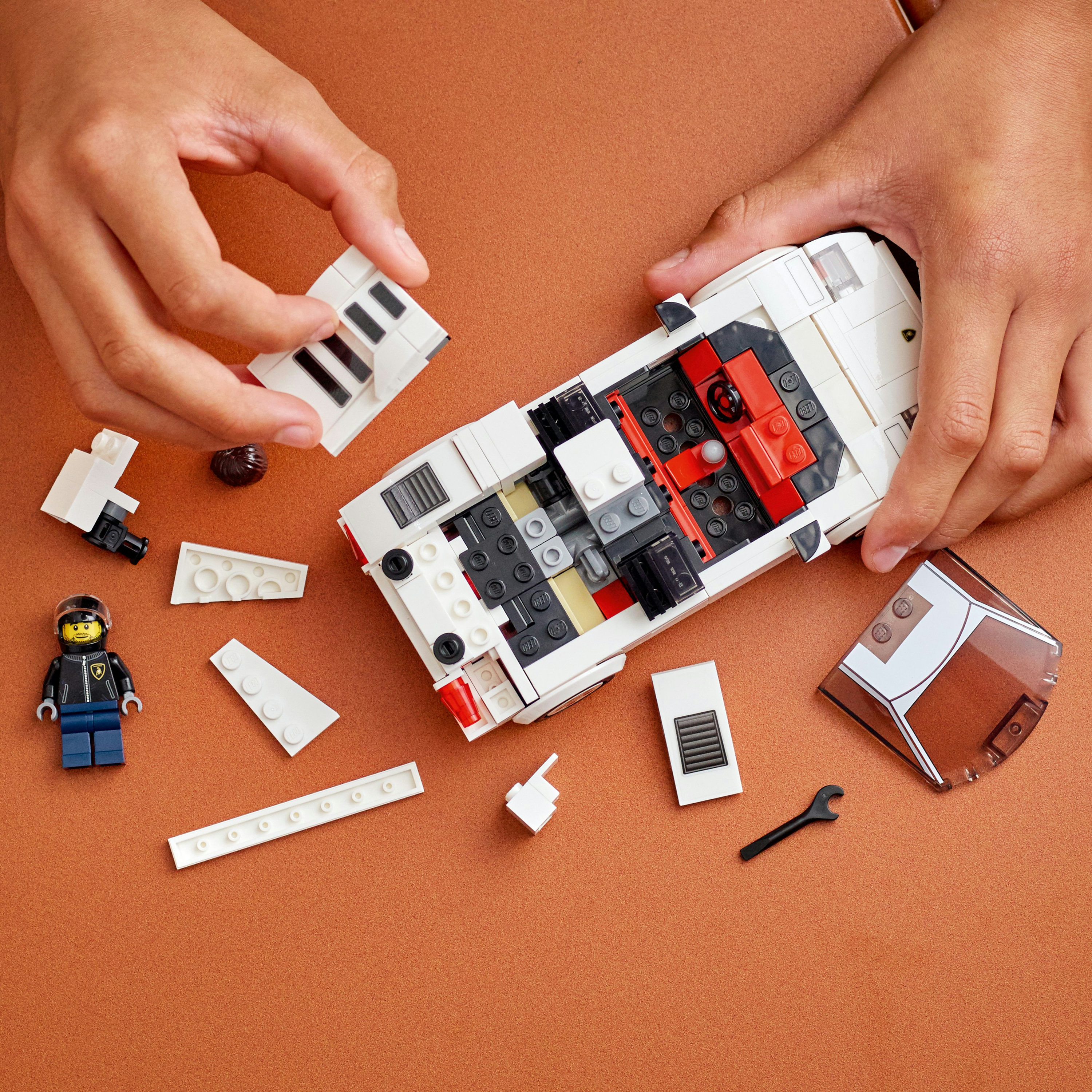 Hände bauen ein weißes Lego-Auto mit schwarzen und roten Details zusammen. Mehrere Teile und eine Minifigur sind in der Nähe.