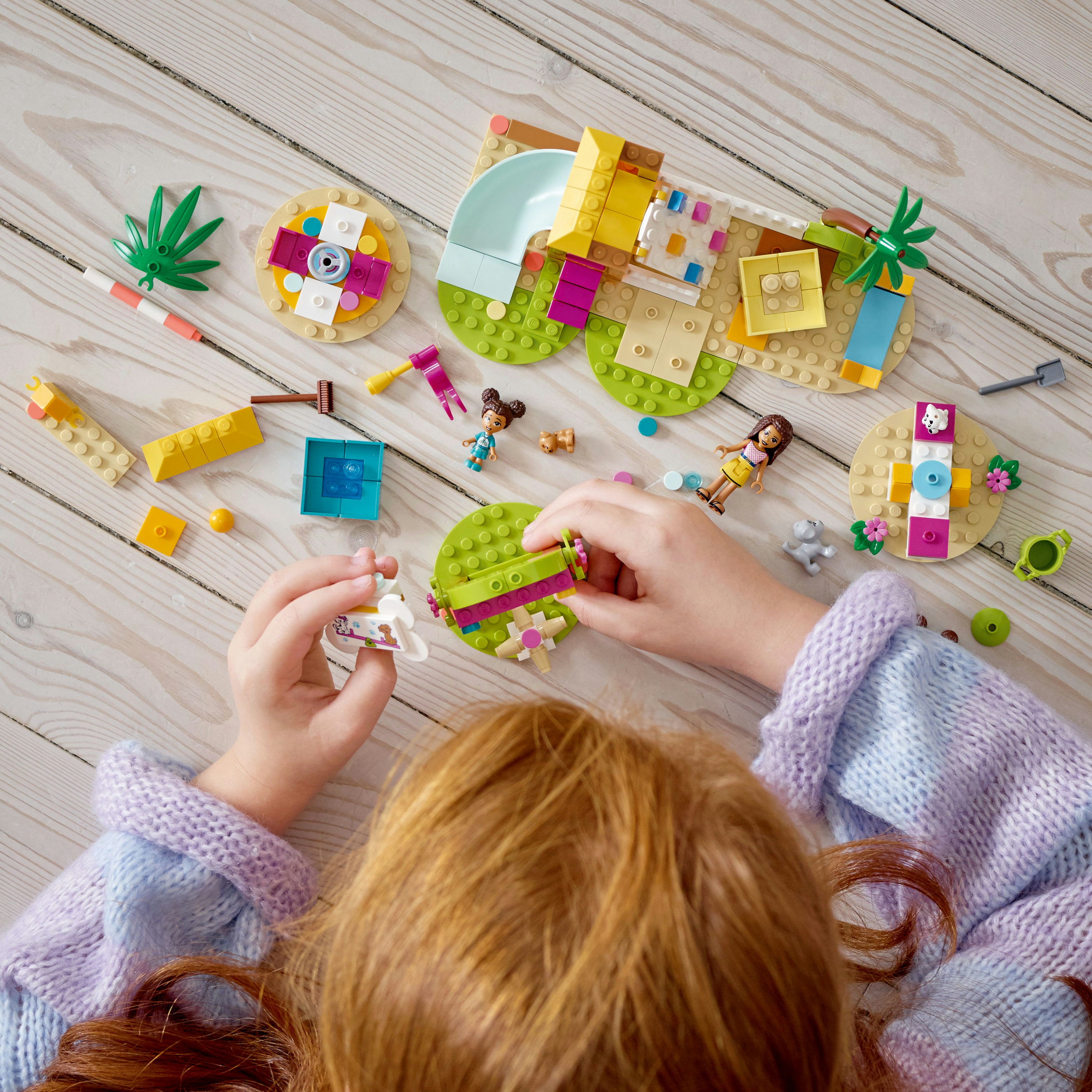 Ein Kind interagiert mit Lego Friends Set, mit verschiedenen Teilen und Figuren auf Holzoberfläche.