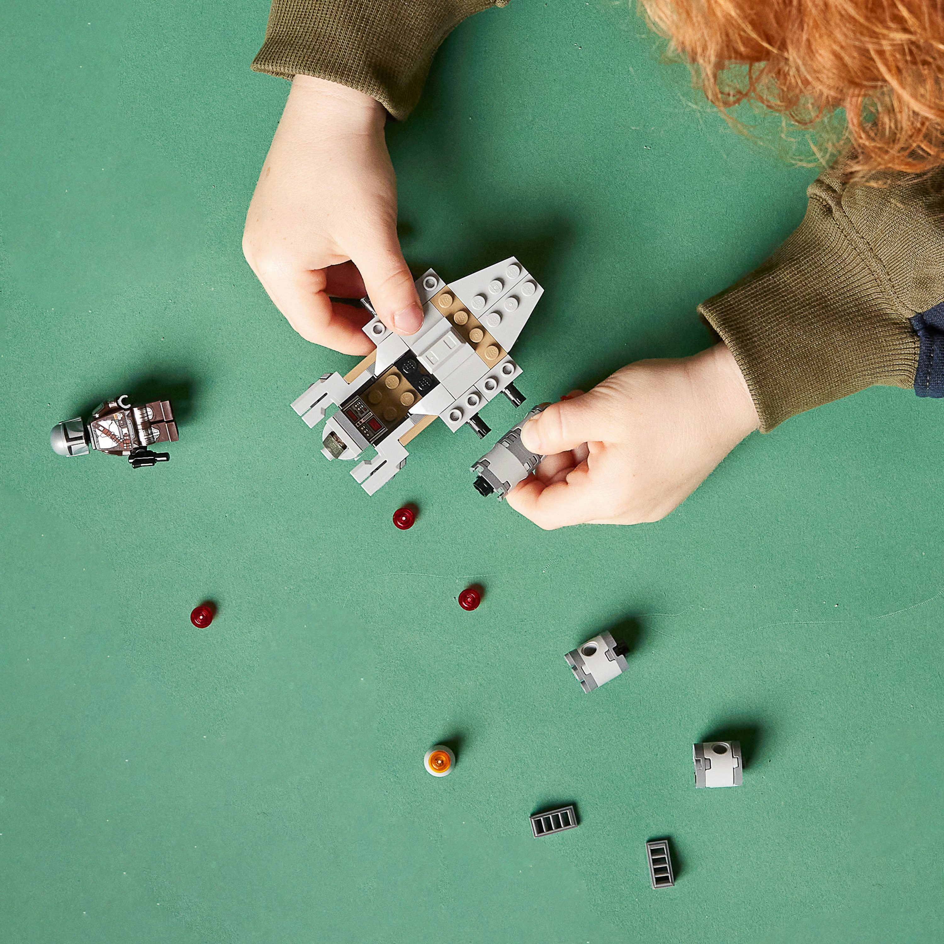 Kinderhände bauen ein graues und beiges Lego-Raumschiff auf grüner Oberfläche, mit verstreuten roten Teilen.