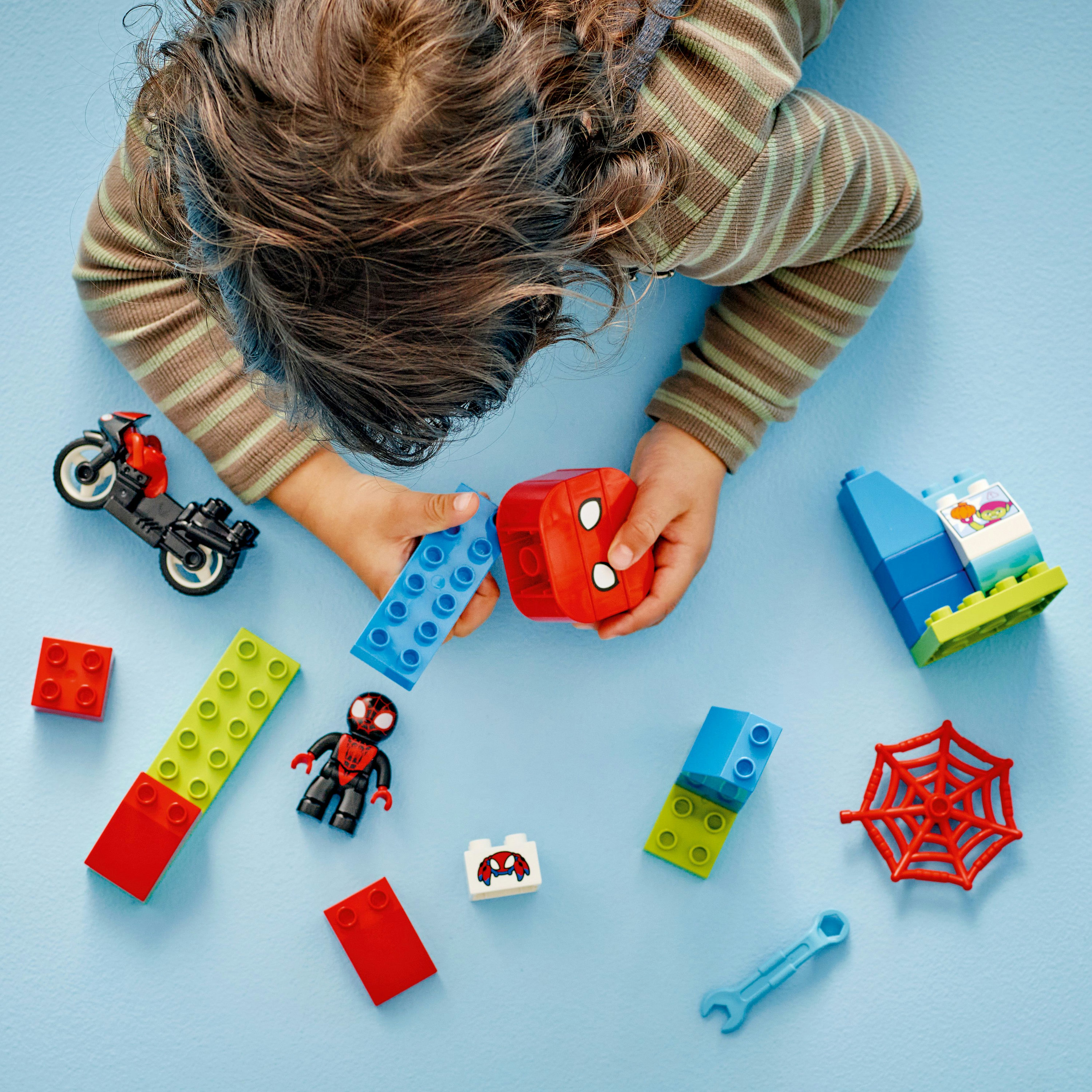 Dziecięce dłonie bawiące się klockami Lego Duplo i figurkami Spider-Mana na niebieskiej powierzchni.