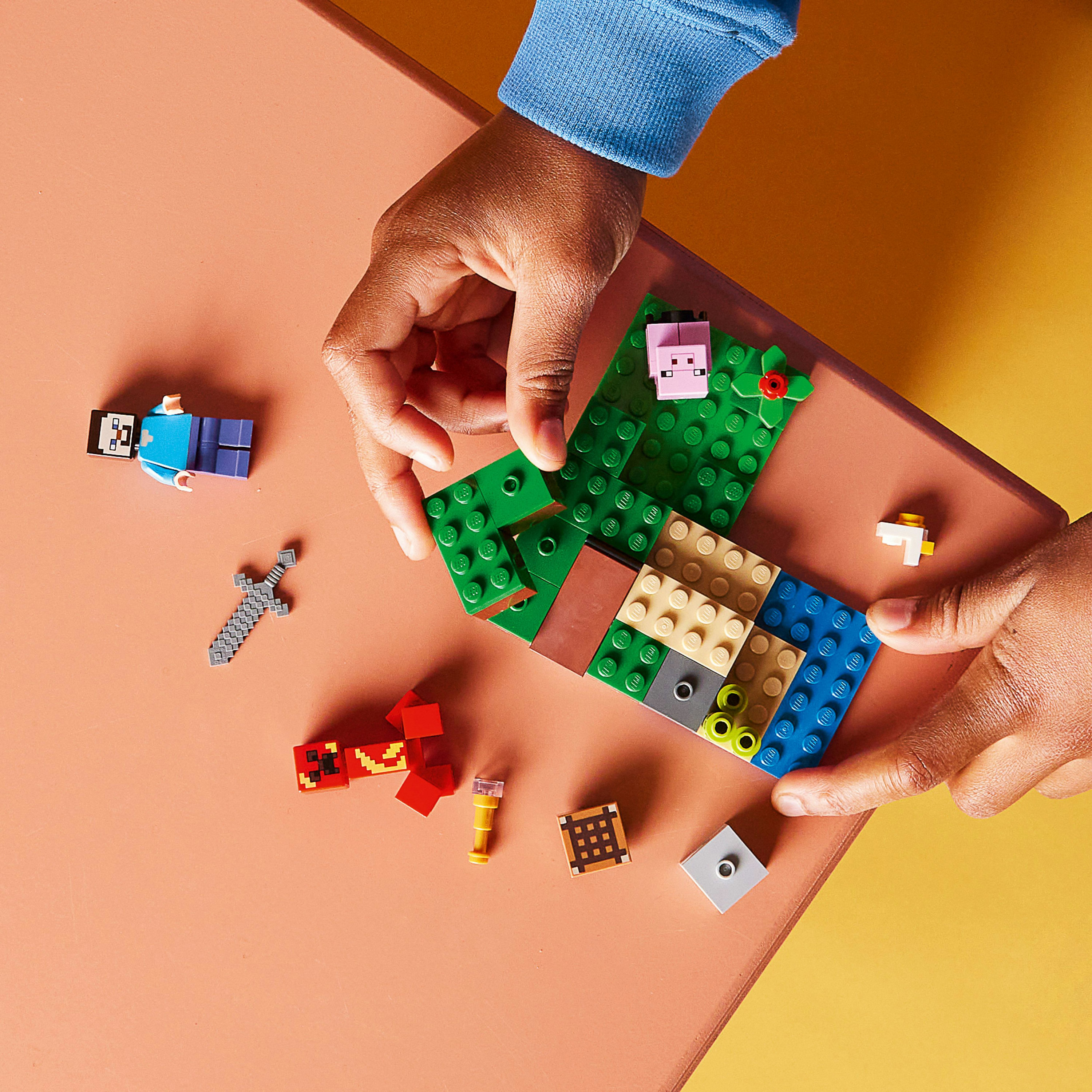 Mani che assemblano pezzi di un set Lego Minecraft su una superficie rosa con mattoncini Lego e minifigure sparse.