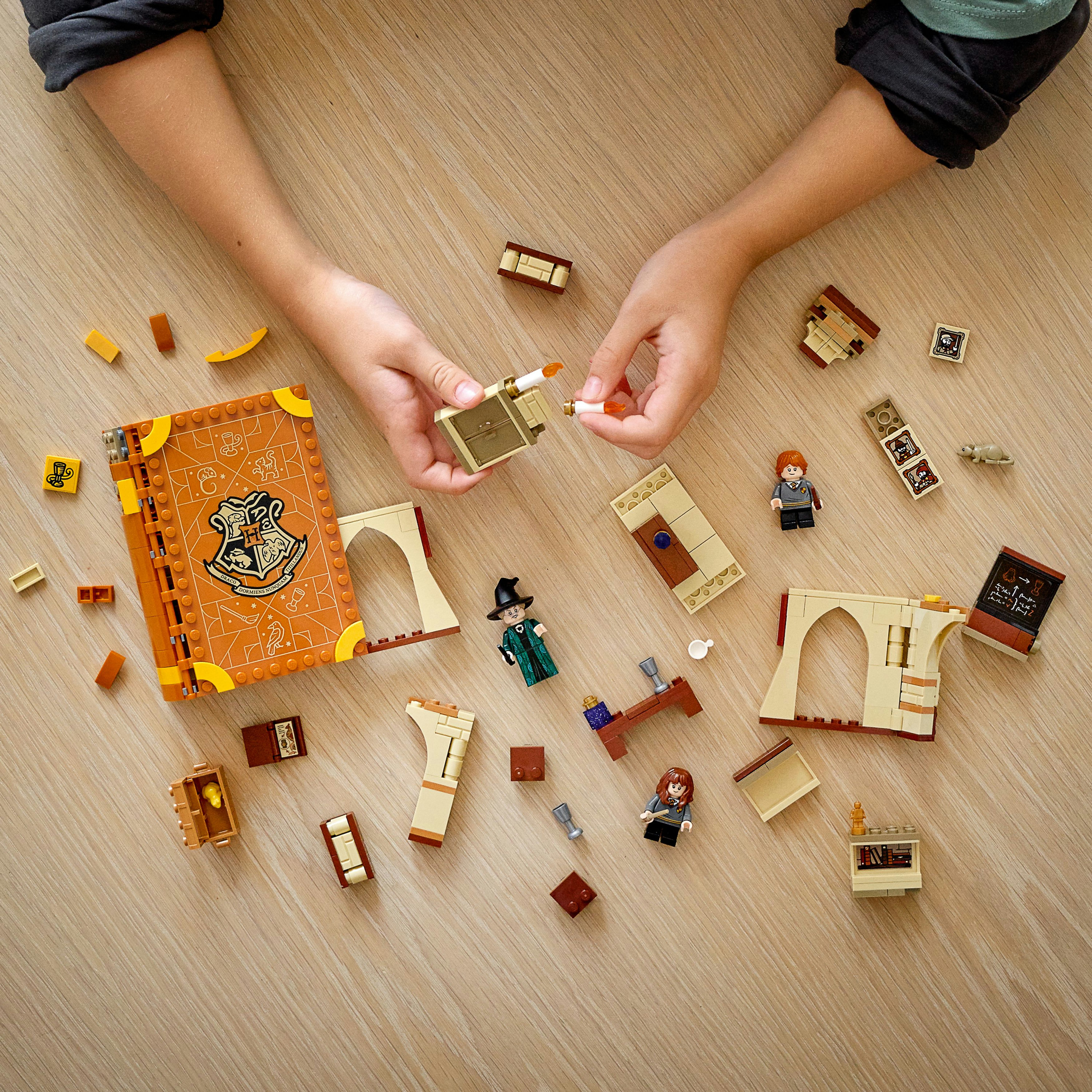 Hände eines Kindes, die LEGO-Teile zusammenbauen, darunter ein Buch und Minifiguren, auf einer Holzoberfläche.