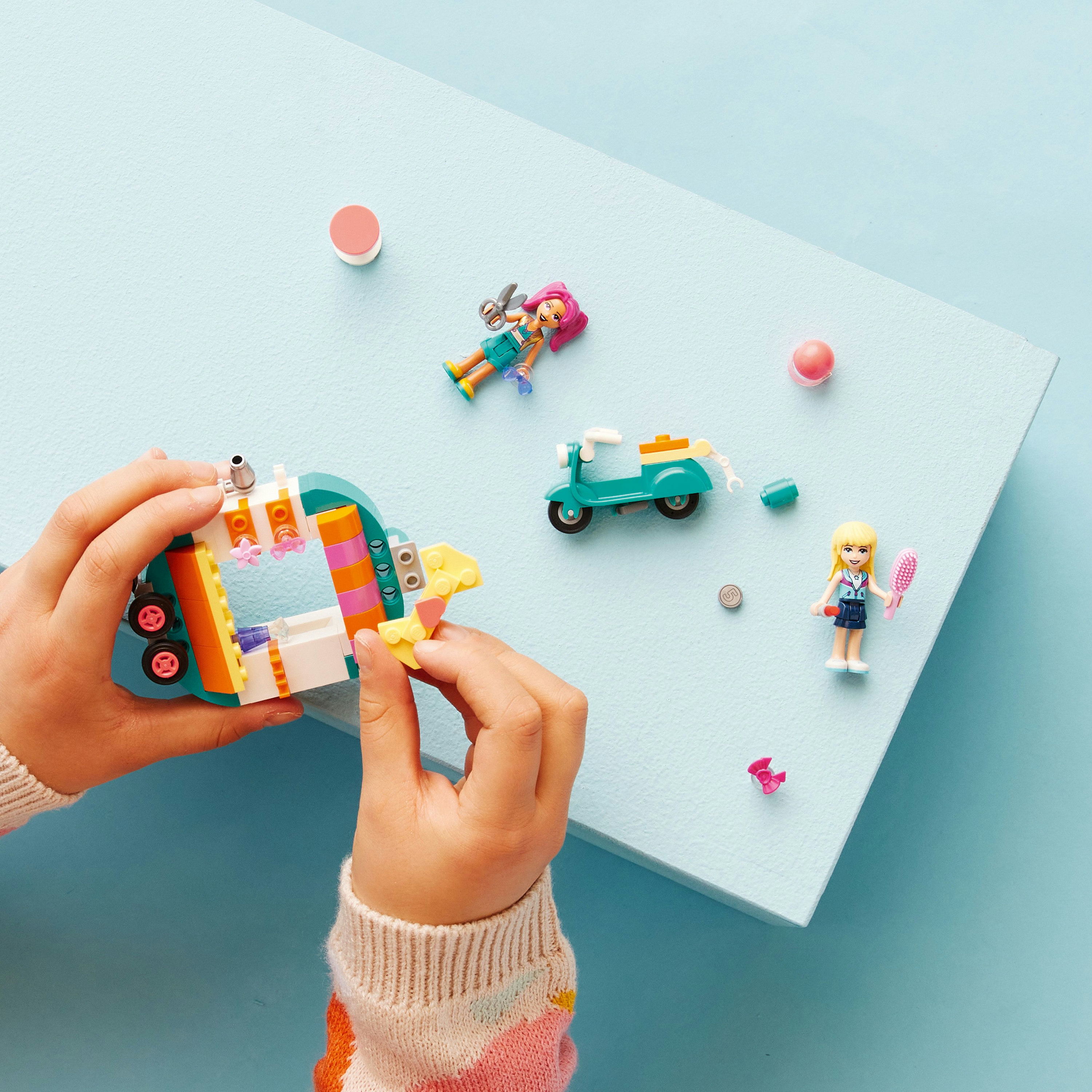 Manos ensamblan un set de Lego con figuras, un scooter y otros elementos sobre una superficie azul claro.