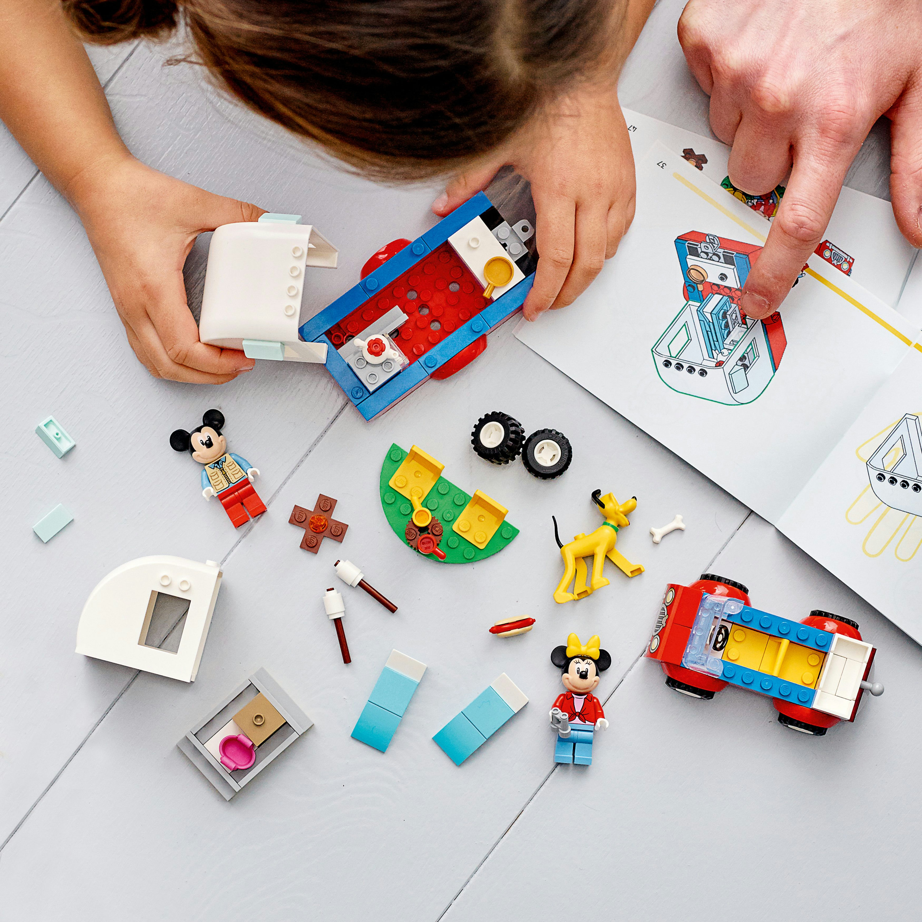Eller, bir araba, figürler ve ahşap bir zeminde talimatlarla Lego setini birleştiriyor.