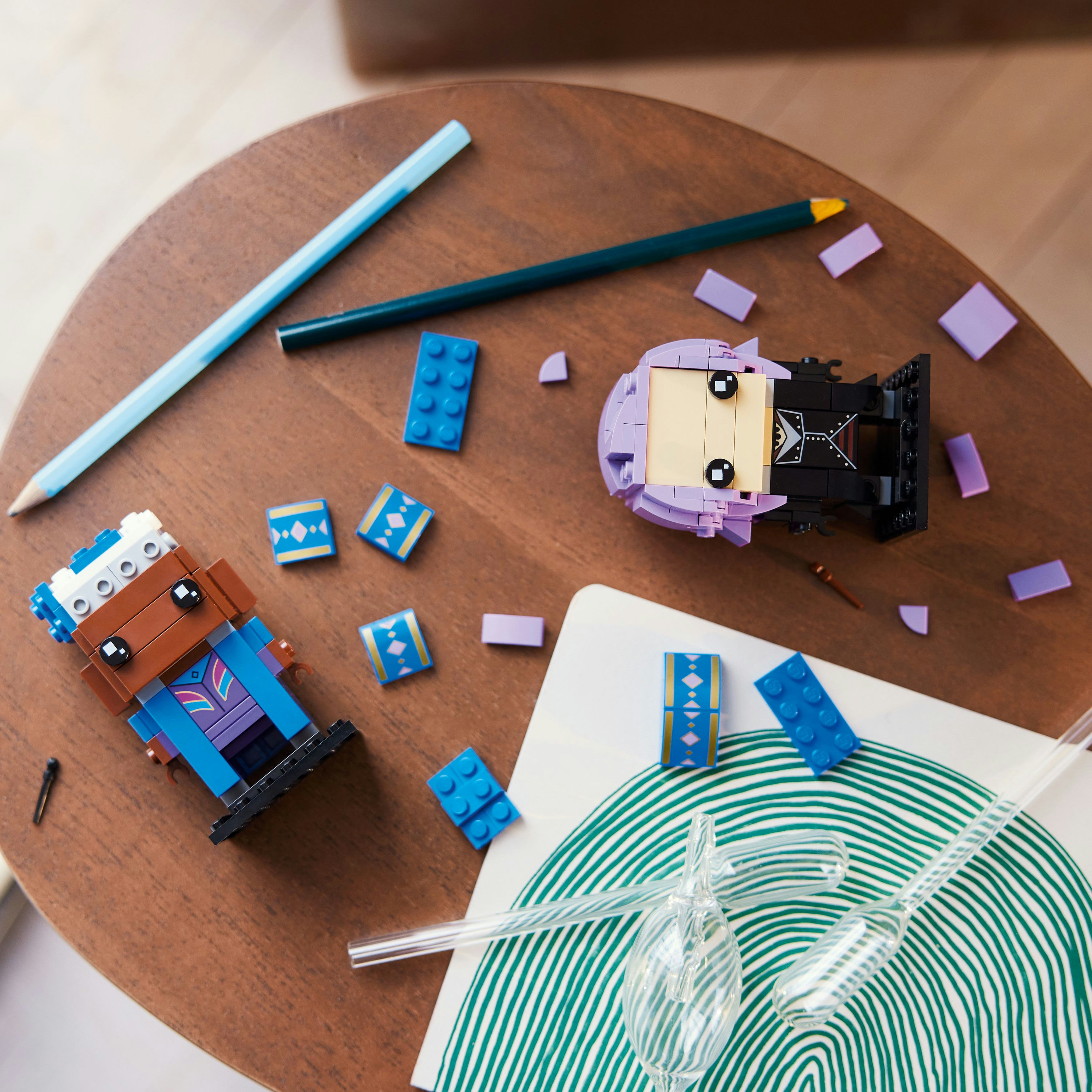 Figurki i elementy LEGO rozrzucone na drewnianym stole. Obecne są również dwa ołówki i notatnik.