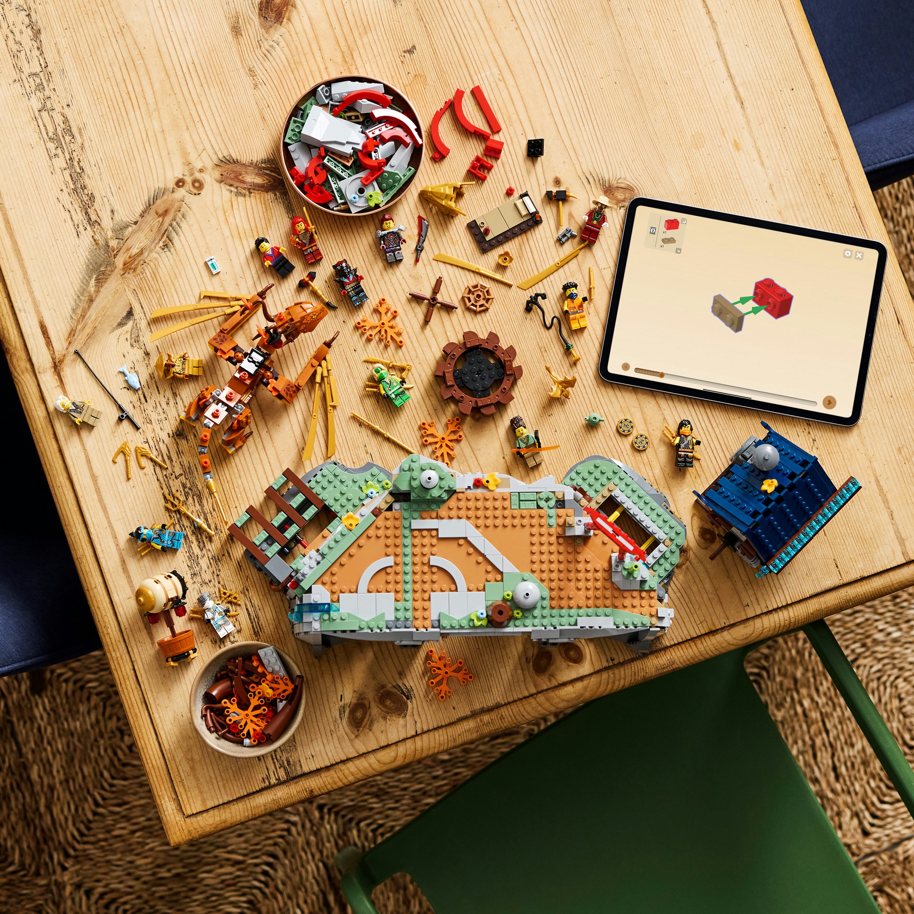 Zestaw Lego z różnymi elementami na drewnianym stole, w tym pojazdem, figurkami i tabletem z instrukcjami.