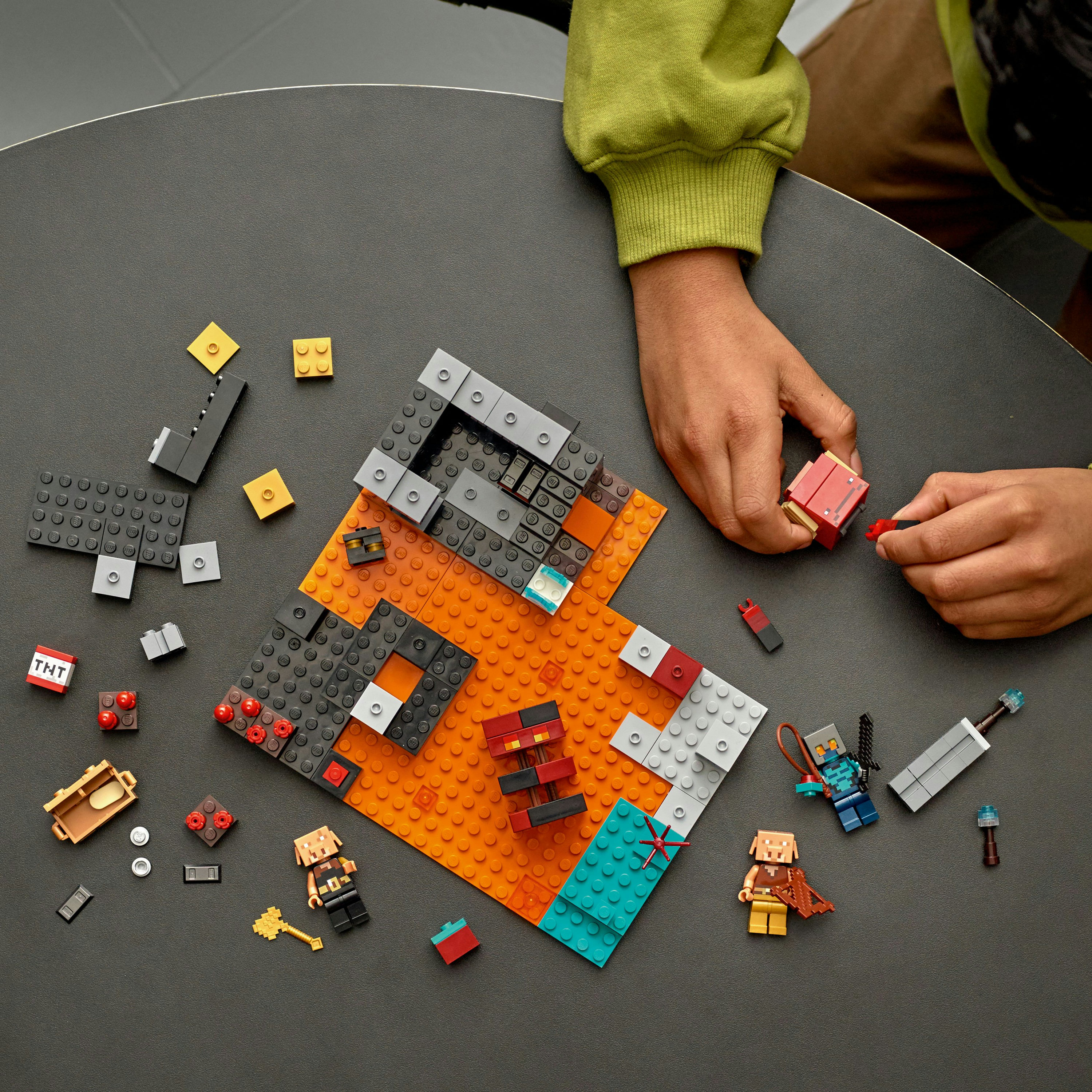 Hände bauen eine Lego Minecraft-Szene mit Figuren, Blöcken und Lava auf einem grauen Tisch.