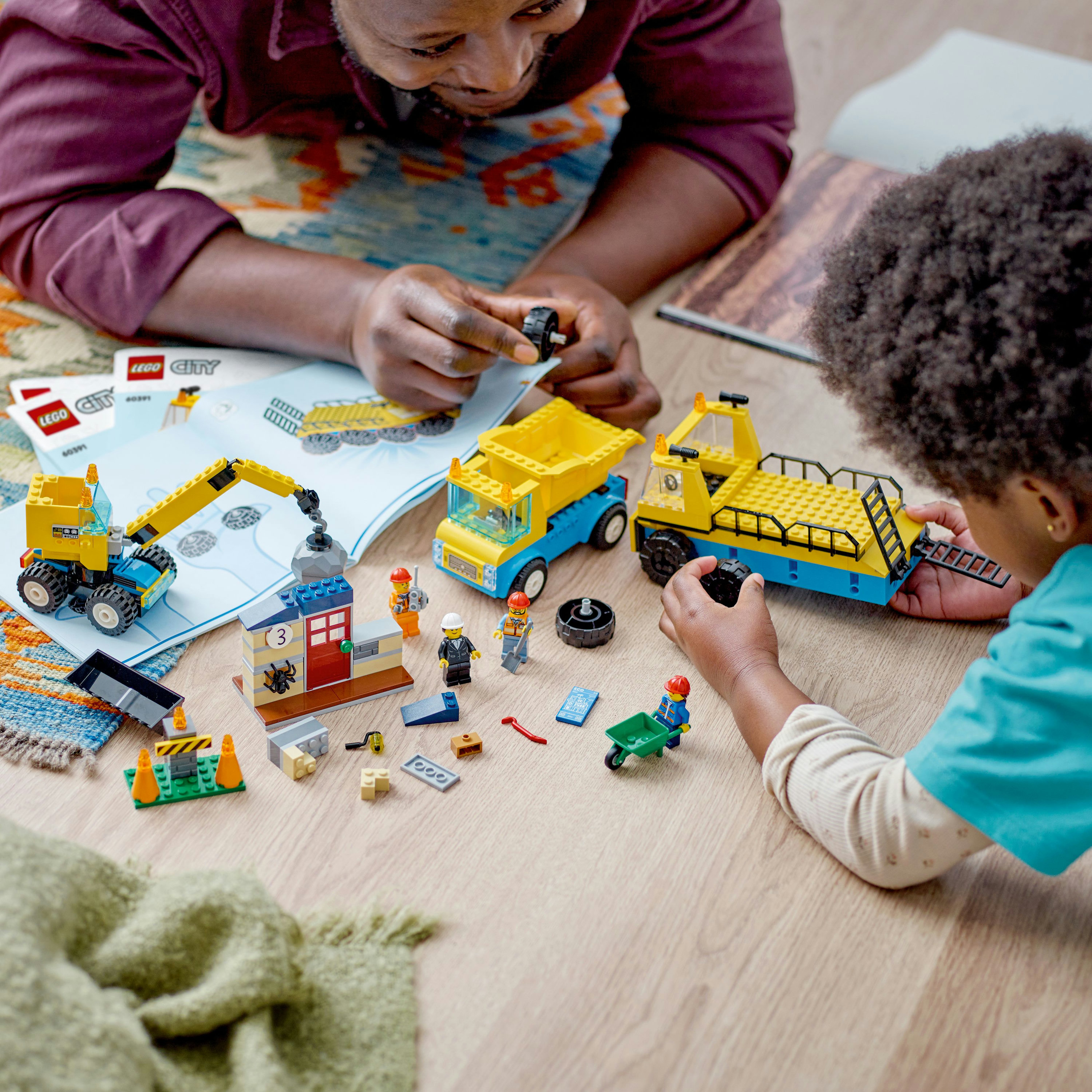 Dos personas ensamblan un set LEGO City en un suelo de madera con camiones, figuras y elementos de construcción.