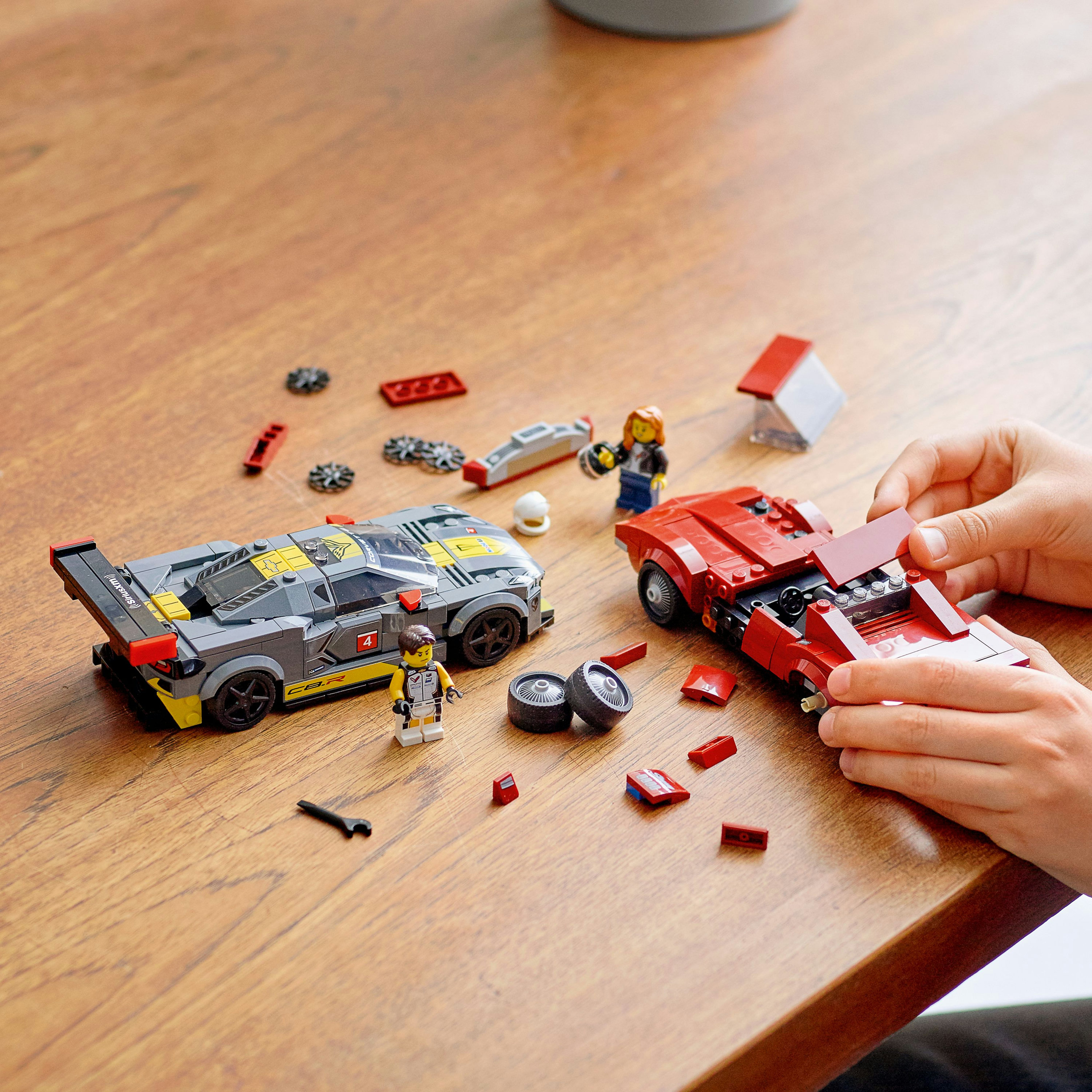 Zwei Lego-Autos werden auf einem Holztisch gebaut, mit Minifiguren und losen Teilen verstreut.