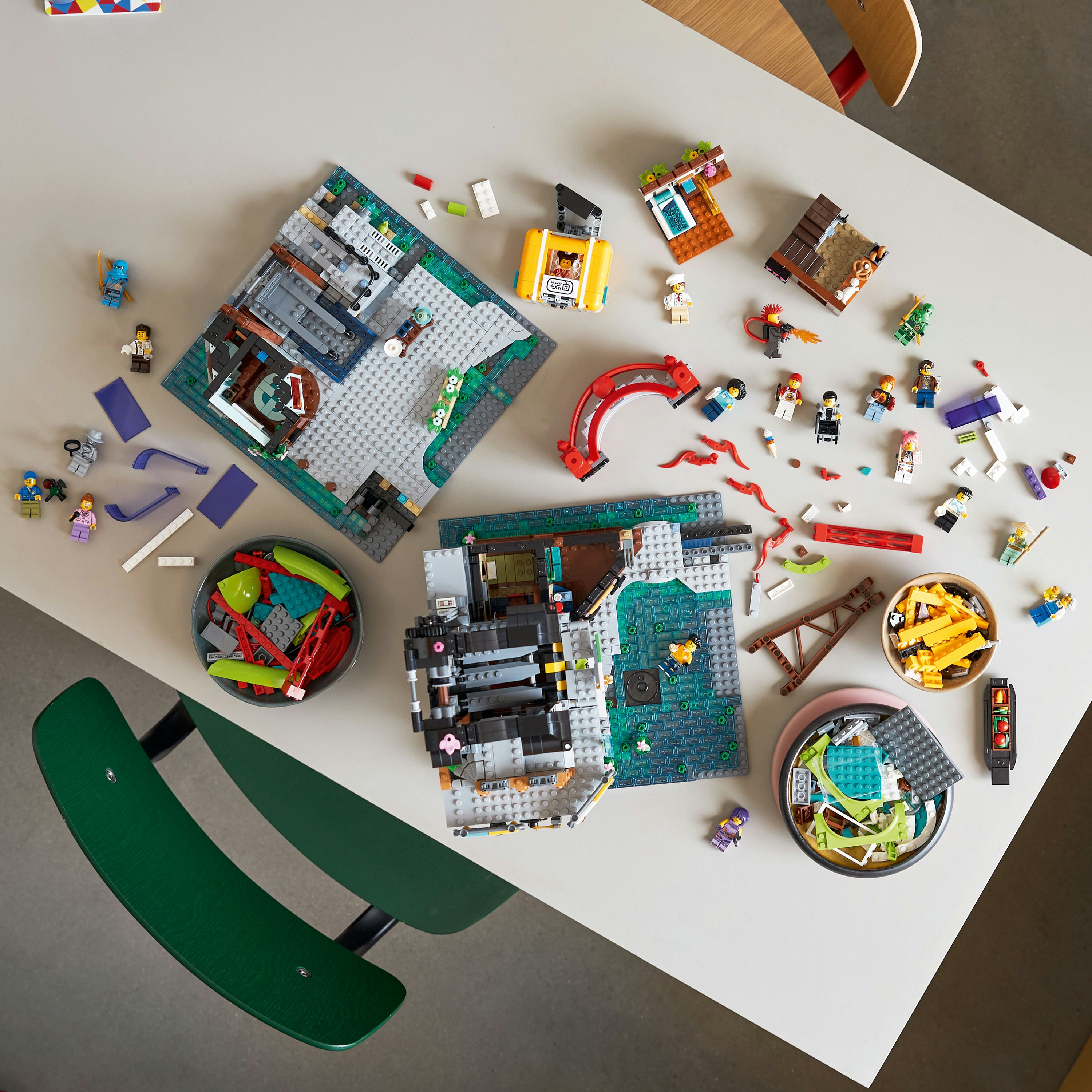 Zestawy LEGO na stole: różne modele, figurki i akcesoria, z zielonymi krzesłami w tle.