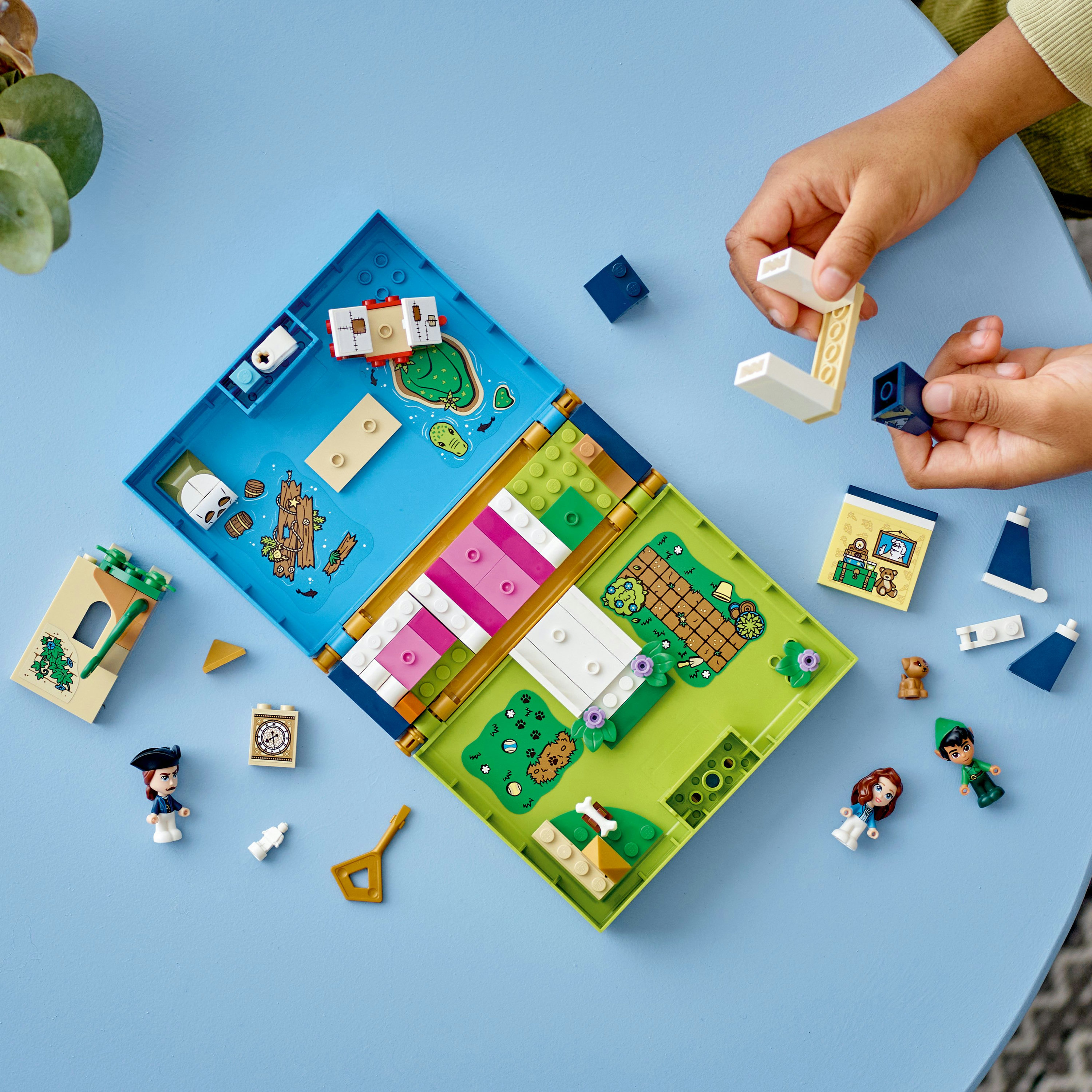 Lego set geopend op blauw oppervlak. Bevat figuren, een huis en een tuin. Handen bouwen.