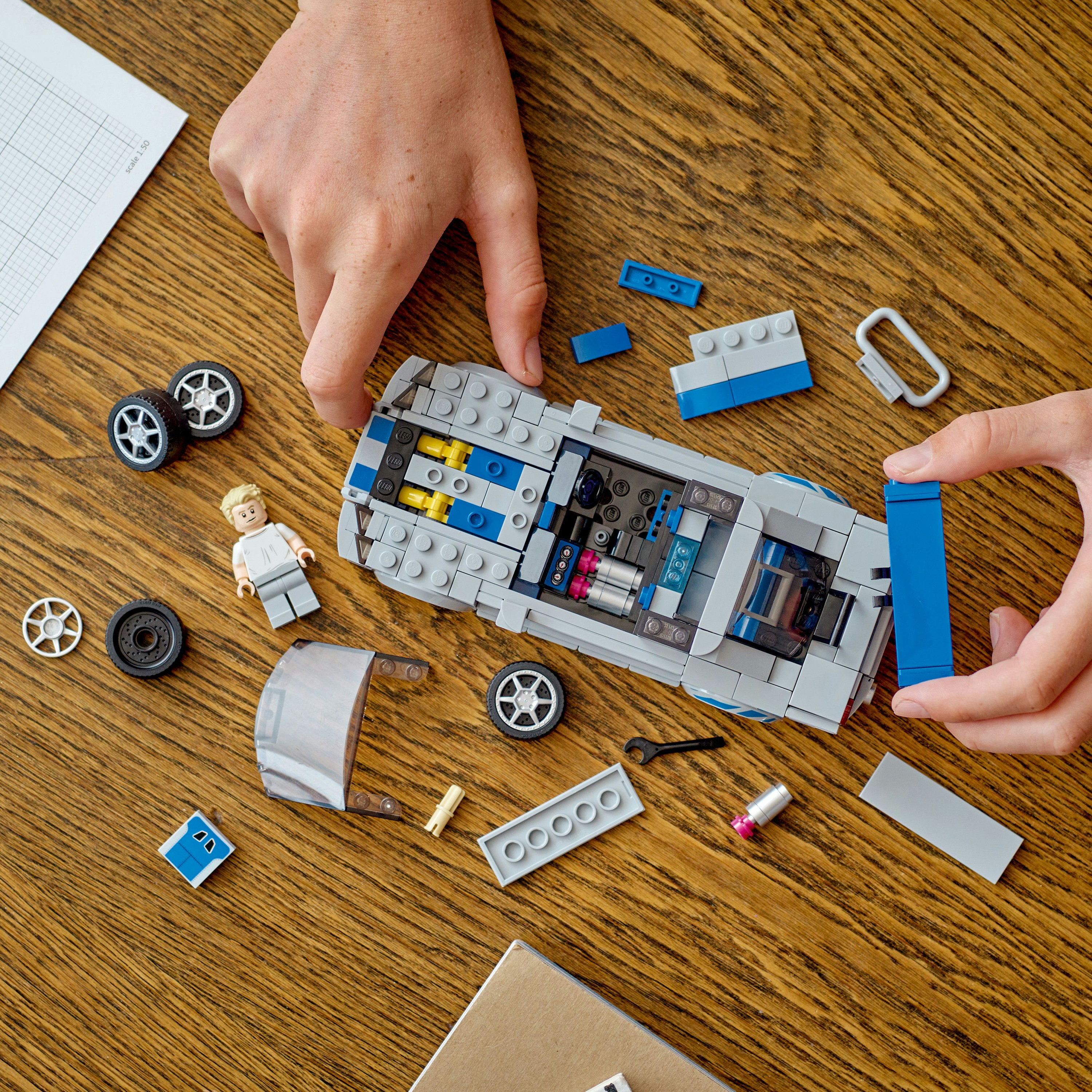Ręce składające samochód Lego, z kołami, miniaturową osobą i dodatkowymi elementami Lego na drewnianej powierzchni.