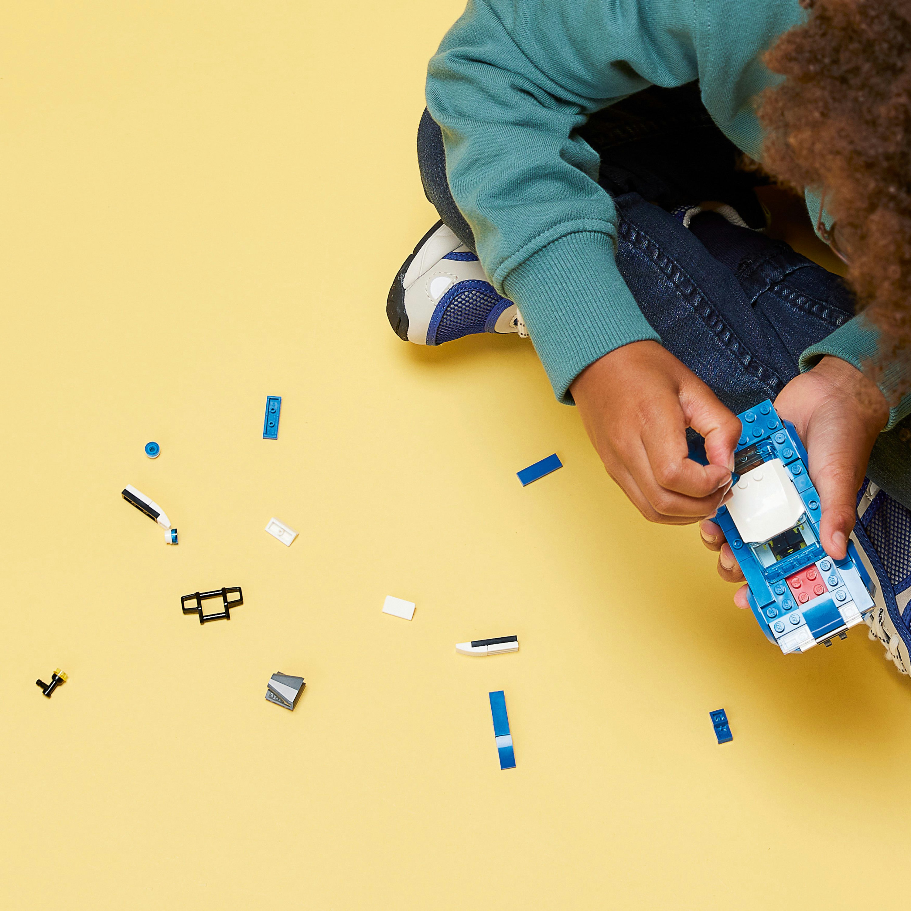 Niño jugando con Lego. Un niño ensambla un coche azul y blanco con piezas sueltas sobre una superficie amarilla.