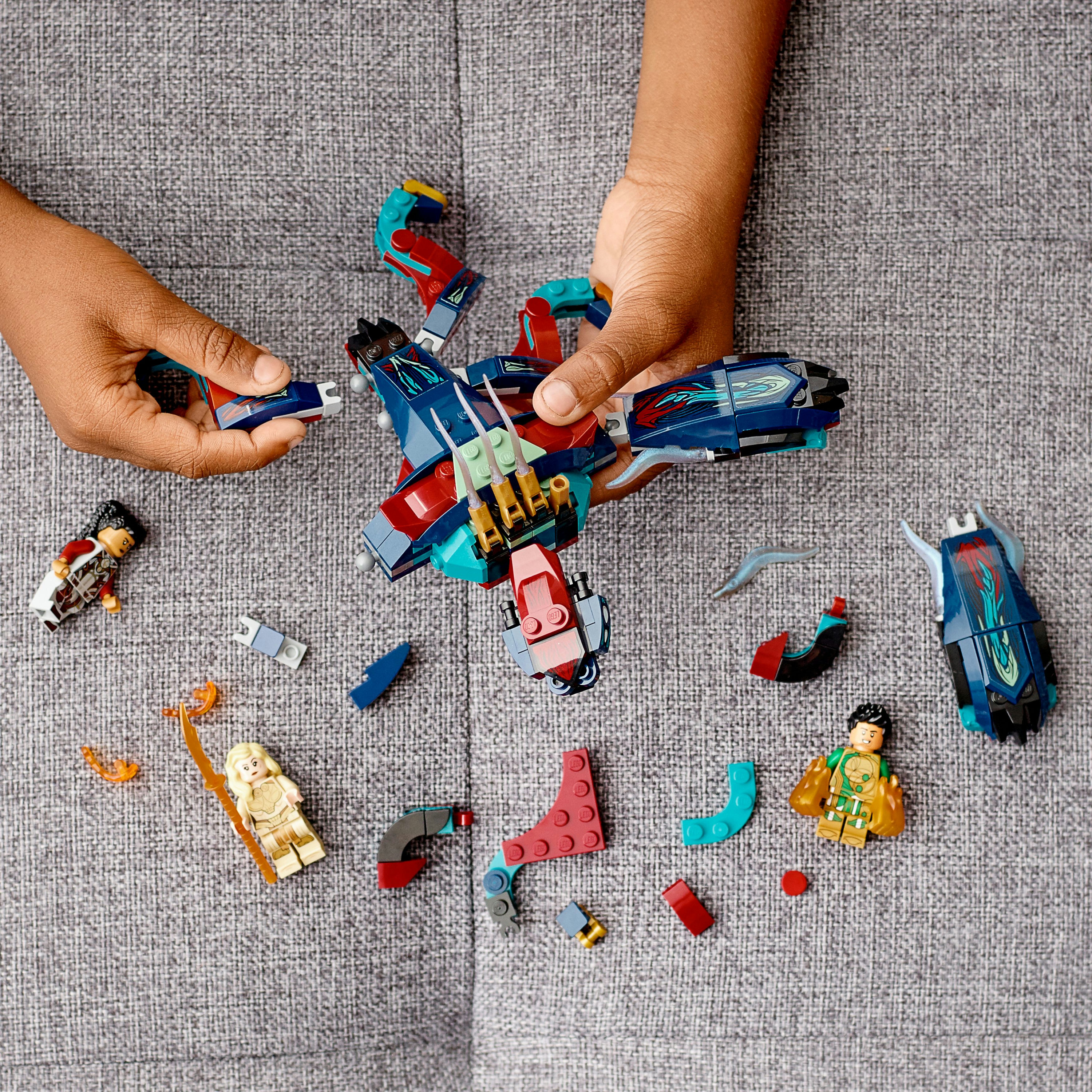 Hände bauen ein Lego-Set mit Figuren und Teilen auf einer grauen Oberfläche zusammen, ein Lego-Flugzeug ist sichtbar.