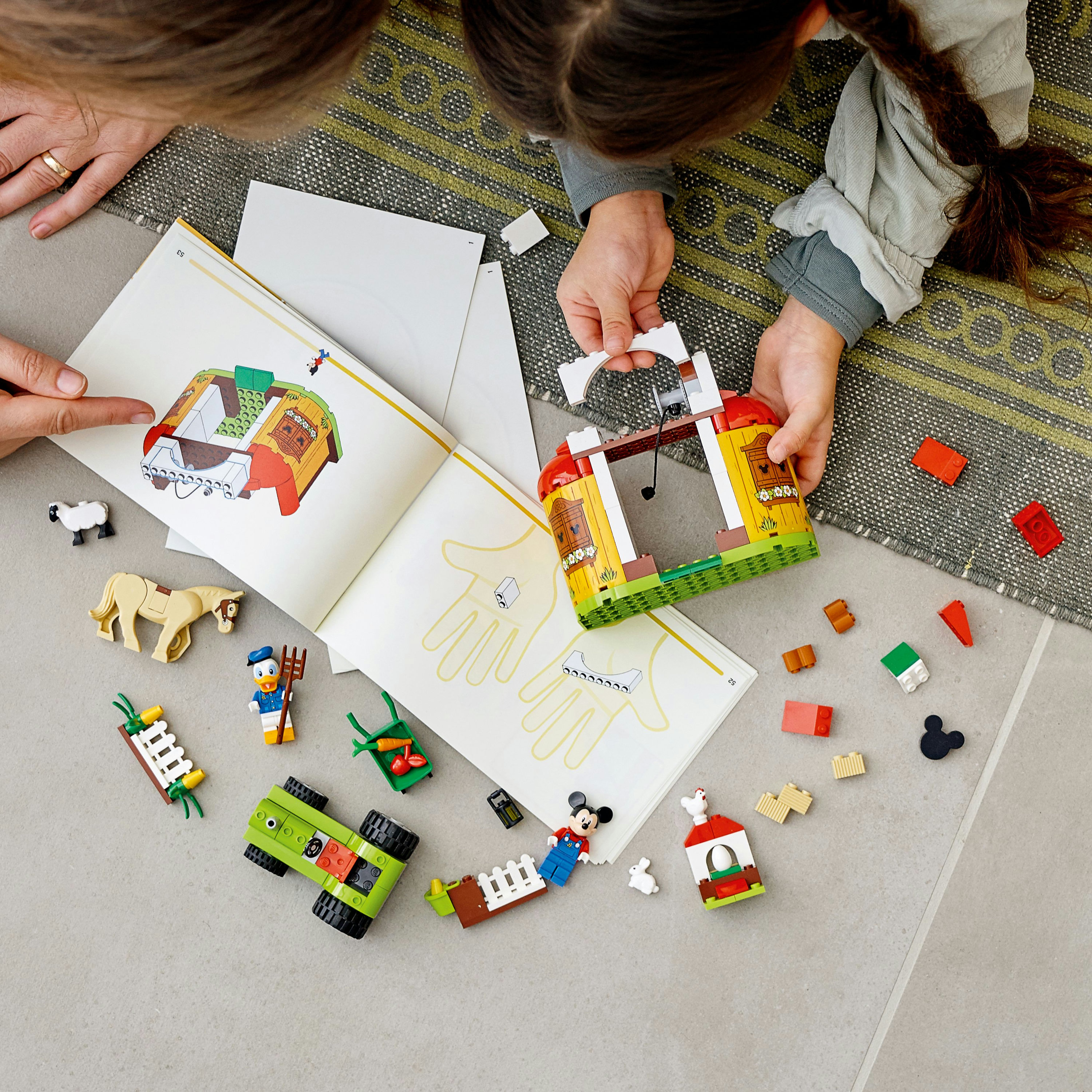 Hände, die eine LEGO Bauernhofszene mit Figuren, Tieren und Bauanleitungen zusammenbauen.