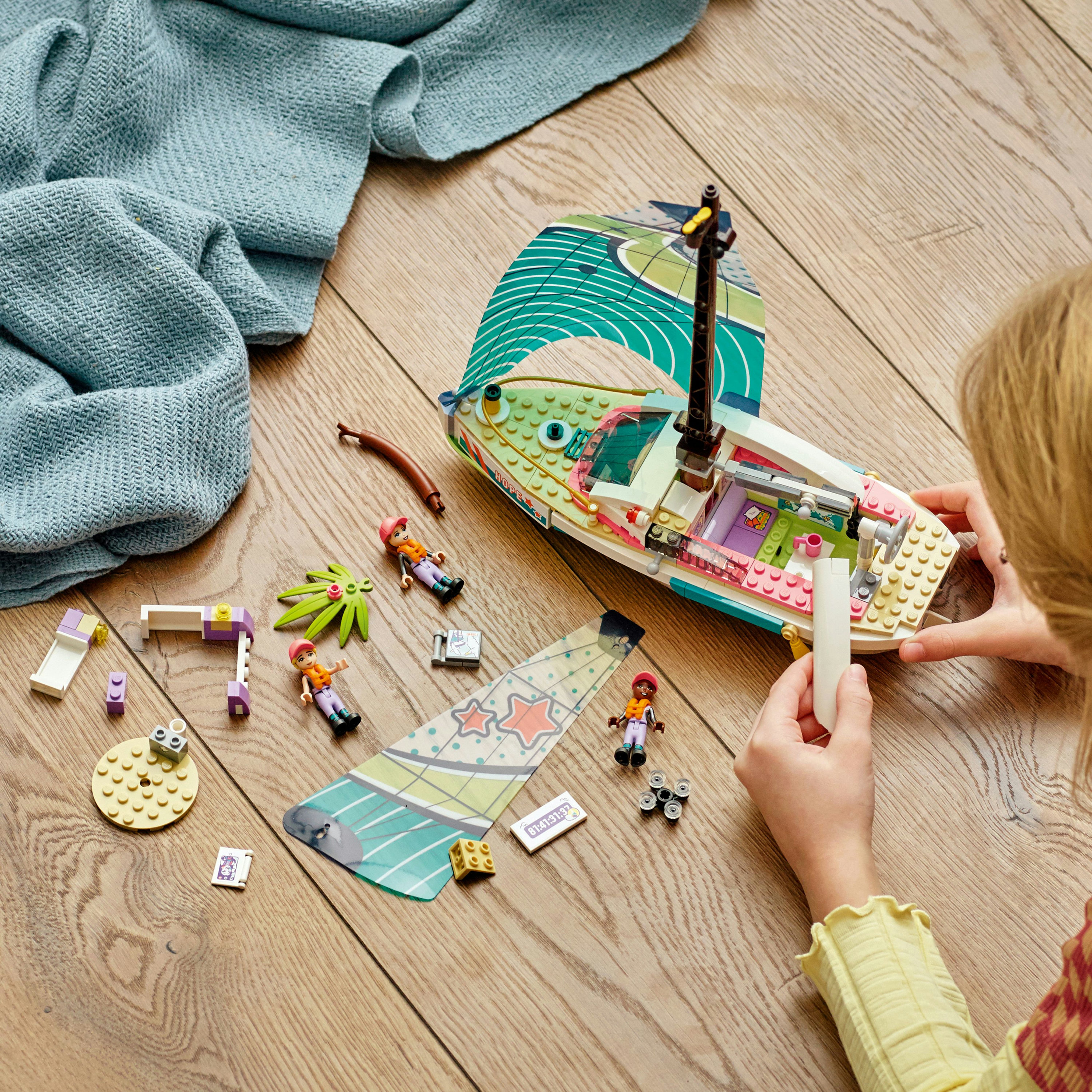 Meisje bouwt een LEGO Friends zeilboot. Boot, minifiguren en diverse accessoires op een houten oppervlak.