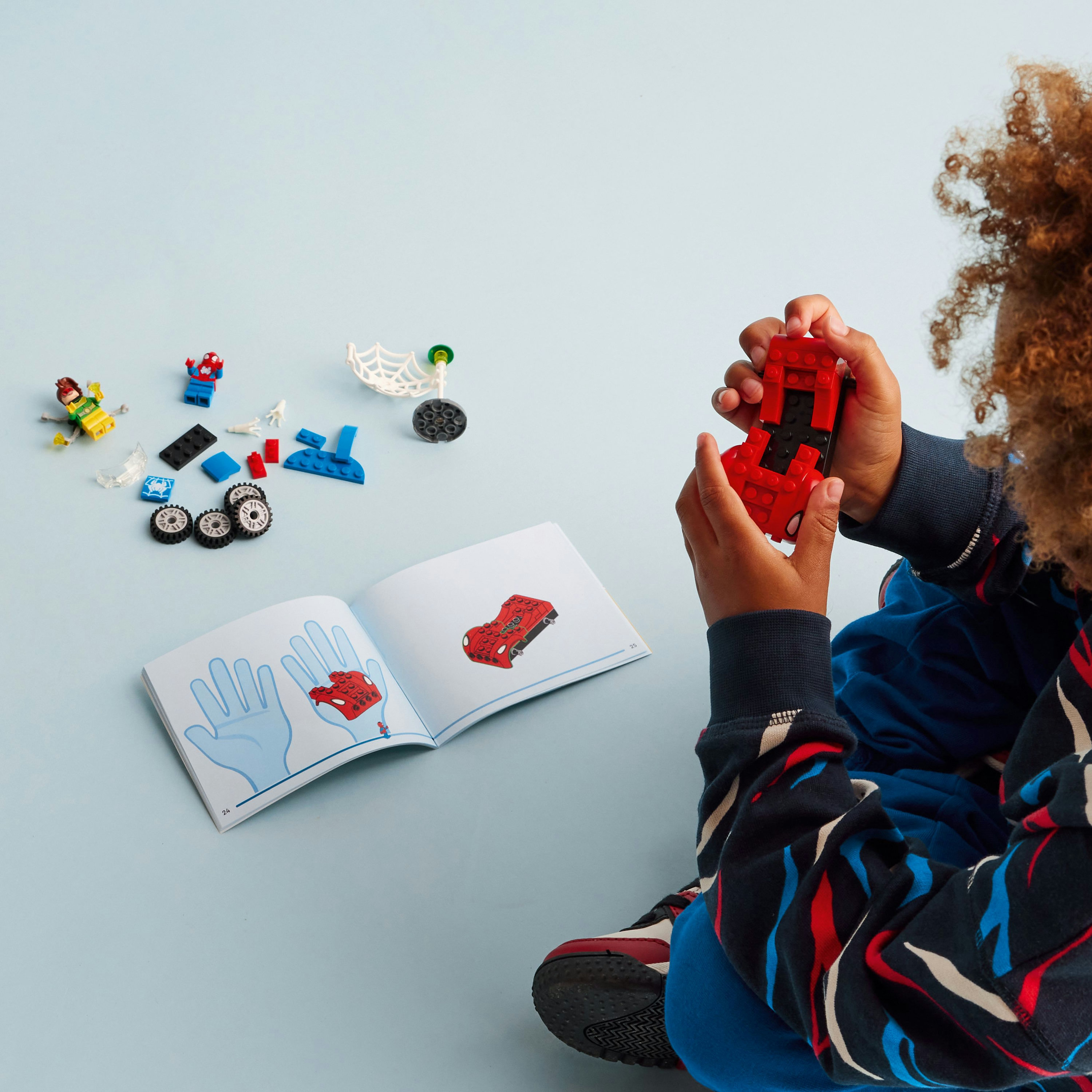 Kind spielt mit Lego. Rotes Spider-Man-Automodell und Anweisungen auf einer blauen Oberfläche mit anderen Lego-Teilen in der Nähe.