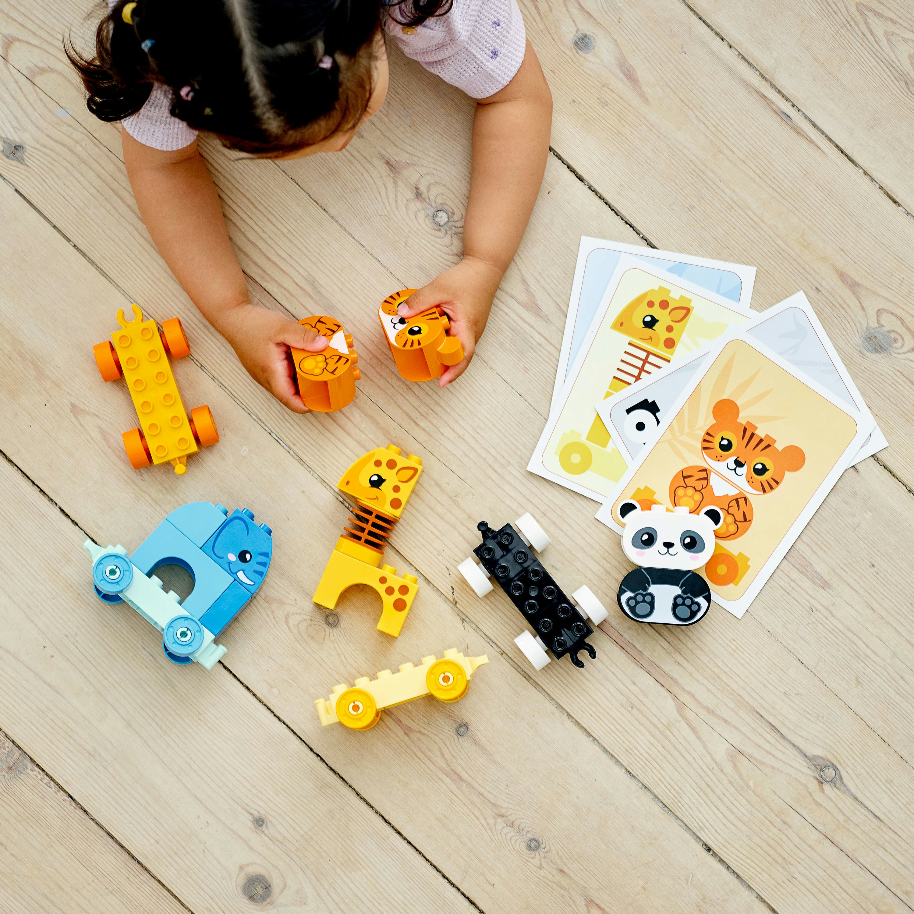 Ein Kind spielt mit Duplo-Spielzeug auf einem Holzboden. Spielzeuge umfassen Tierfiguren und Autos. Karten sind ebenfalls vorhanden.