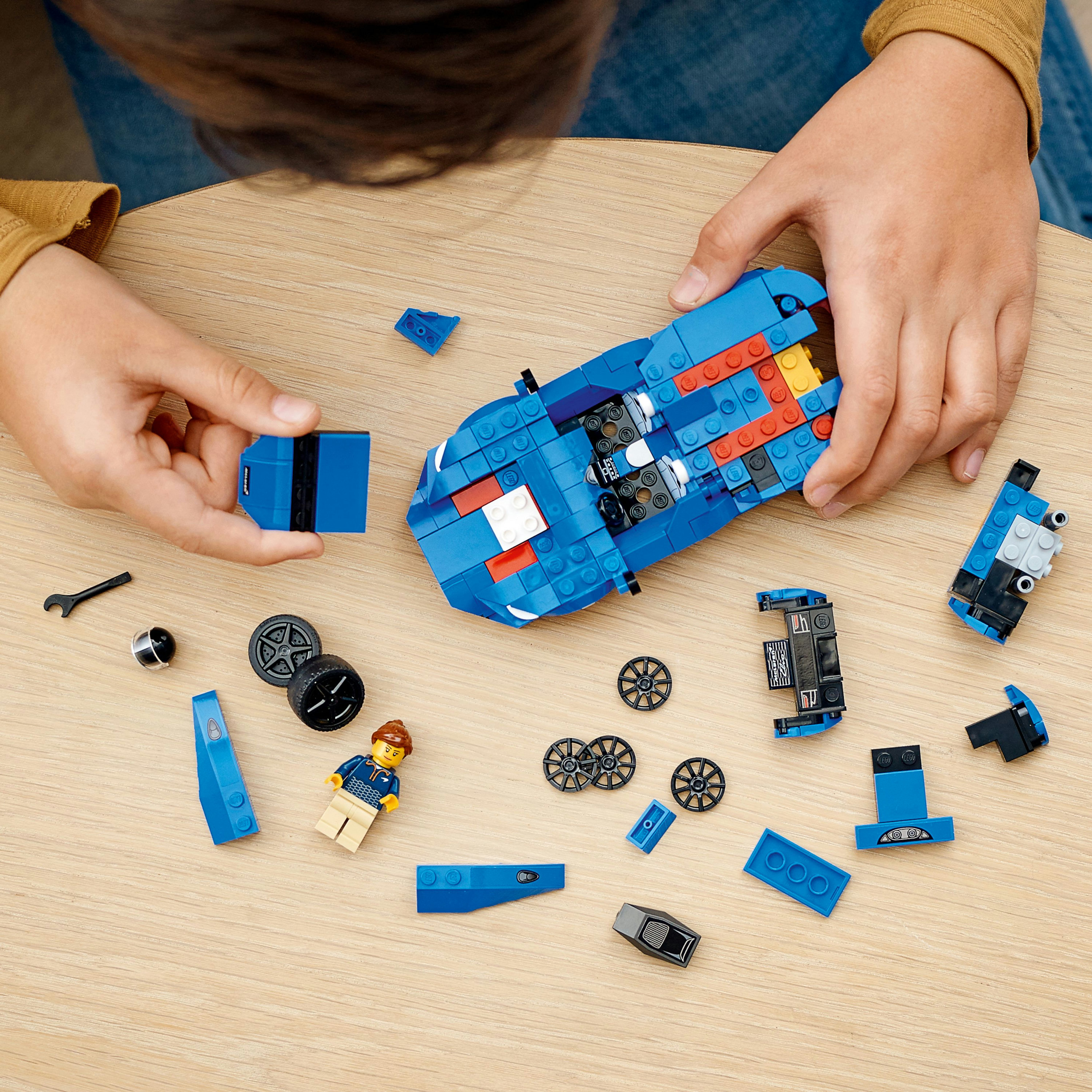 Eller, ahşap bir yüzeyde çeşitli parçalar ve bir minifigür ile mavi bir Lego araba topluyor.