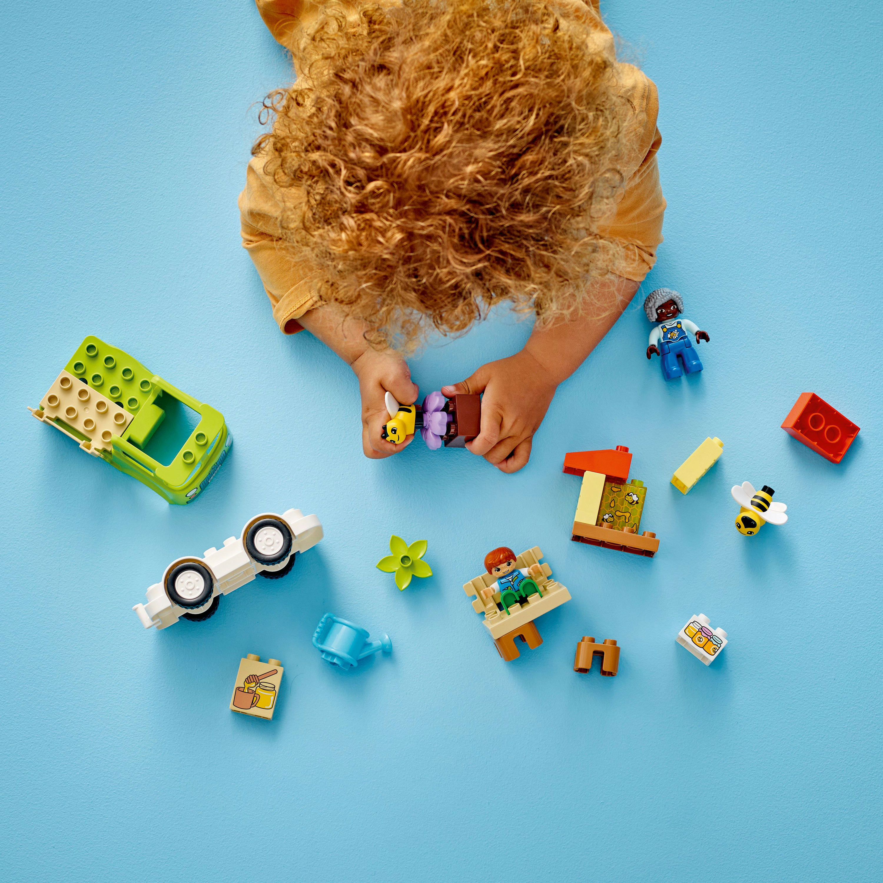 Dziecko patrzy na klocki Lego, w tym zielony samochód, pszczołę, ławkę i inne kolorowe klocki na niebieskiej powierzchni.