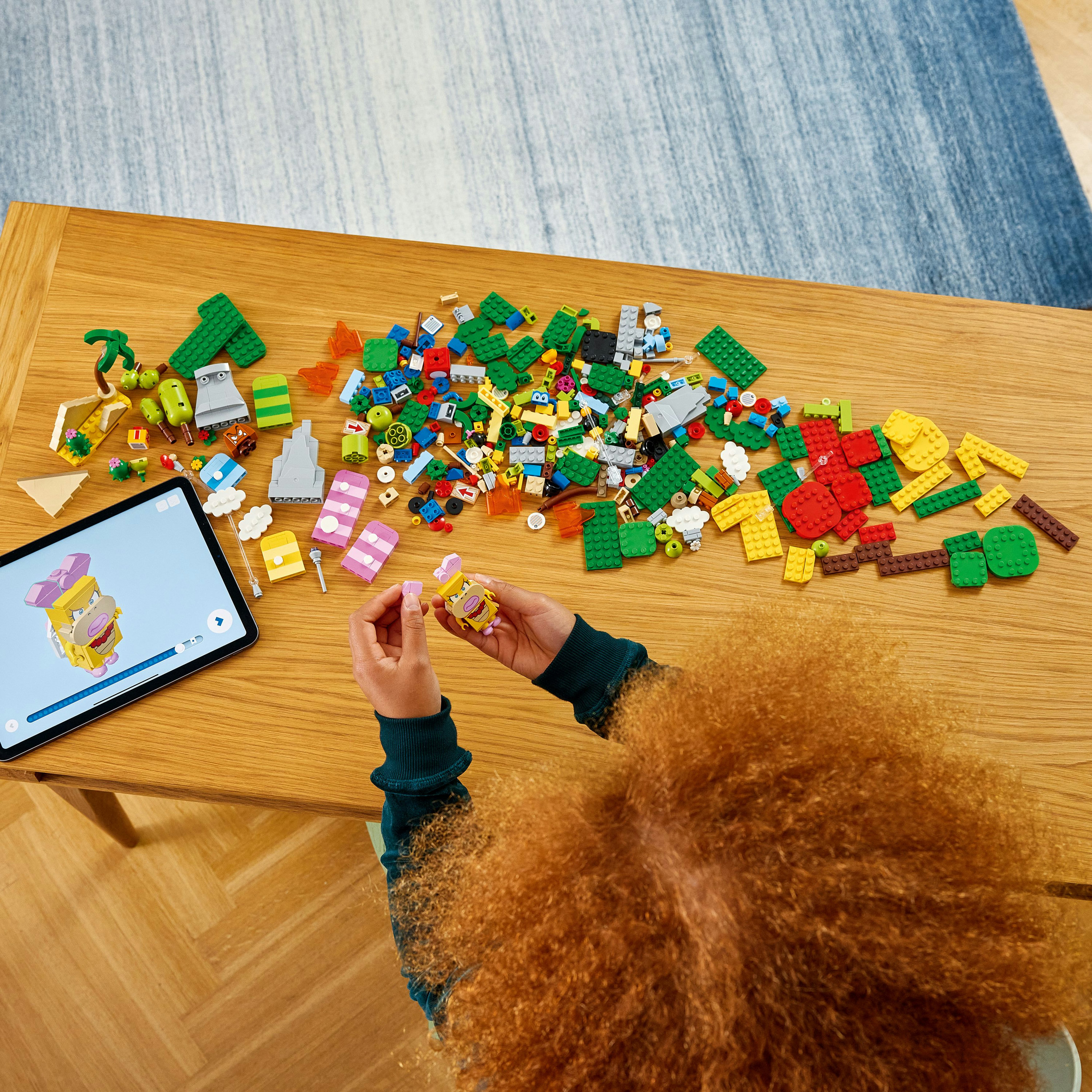 Een persoon met krullend rood haar bouwt een Lego-set op een houten tafel met een tablet.