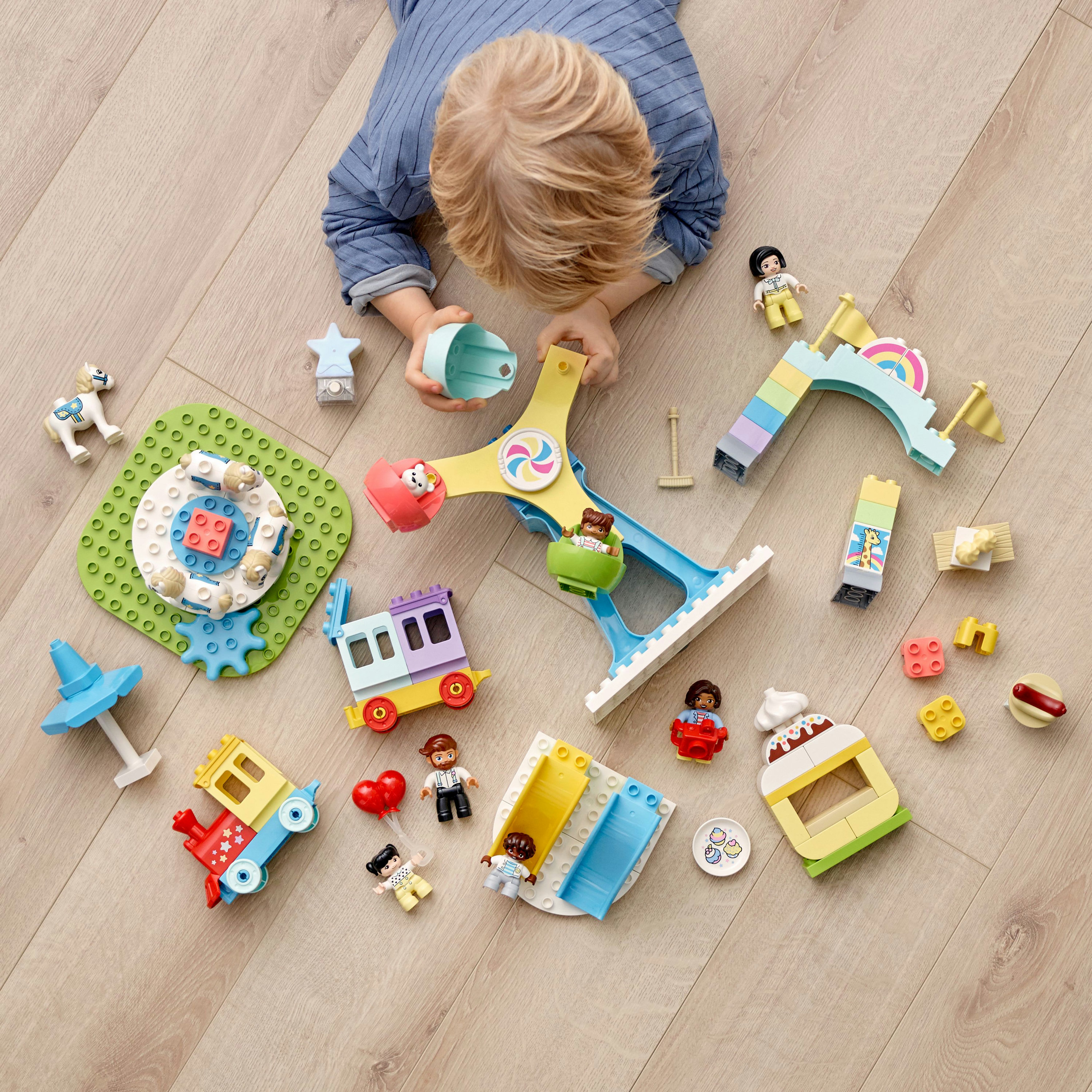 Ein Kind spielt mit Duplo-Blöcken und baut eine Jahrmarktszene mit Karussell, Zug und Figuren auf einem Holzboden.