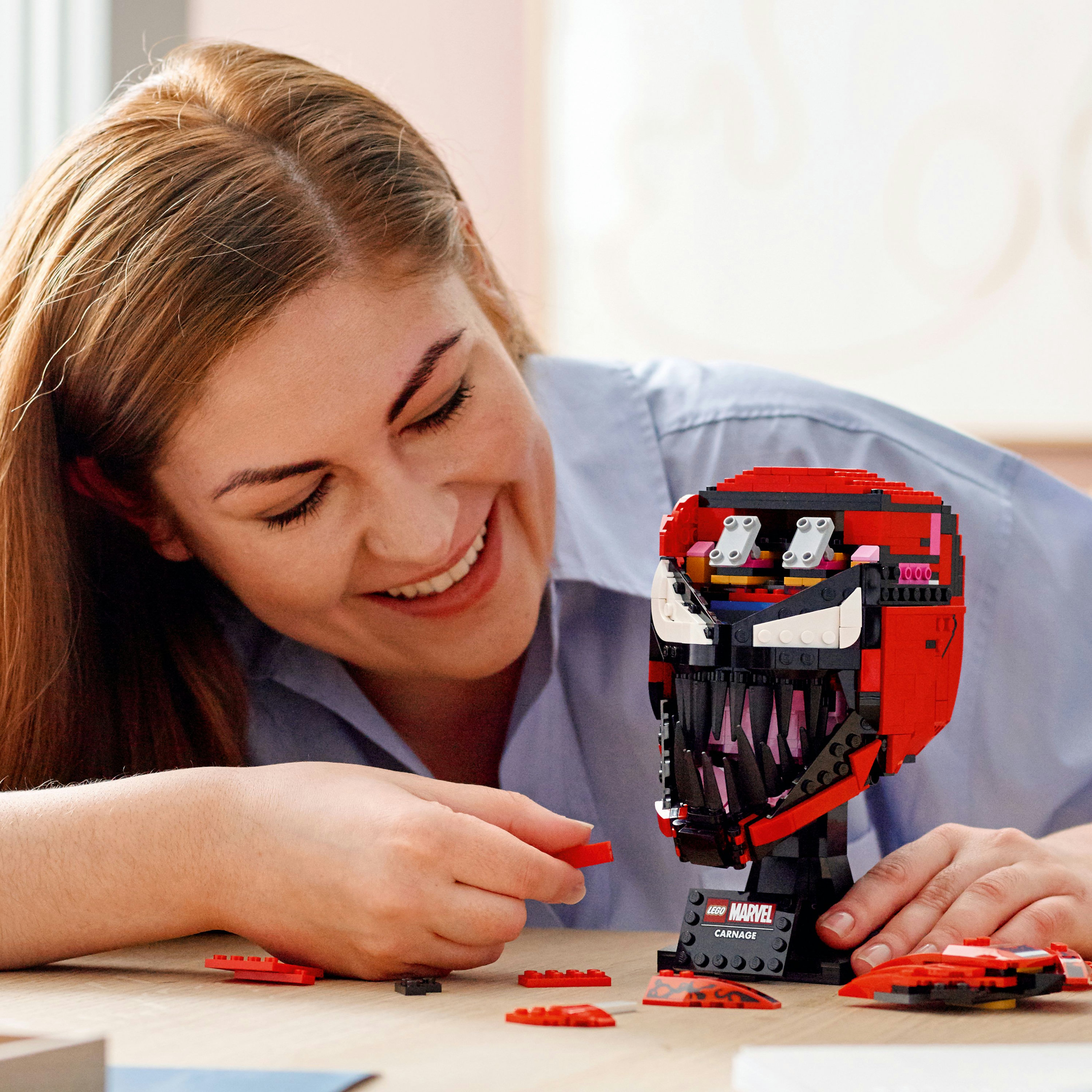 Donna che costruisce il busto LEGO Carnage. Colori rosso e nero. Mattoncini LEGO e istruzioni visibili.