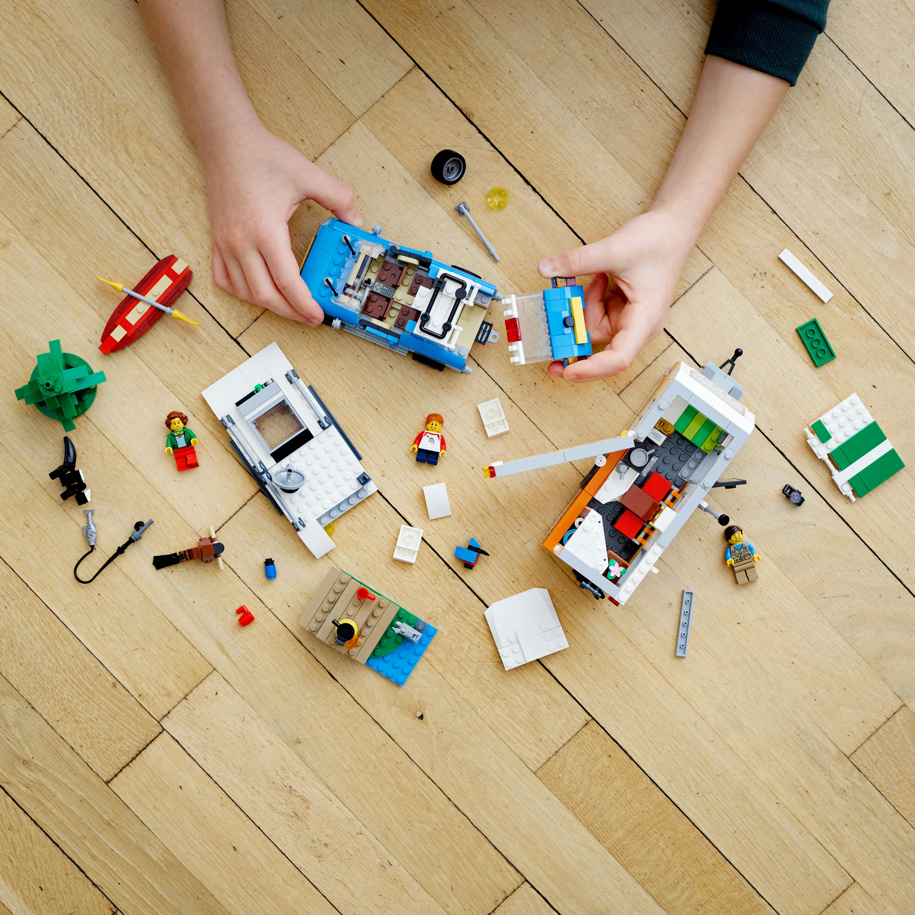 Hände bauen Lego-Fahrzeuge, Teile liegen auf einem Holzboden verstreut.