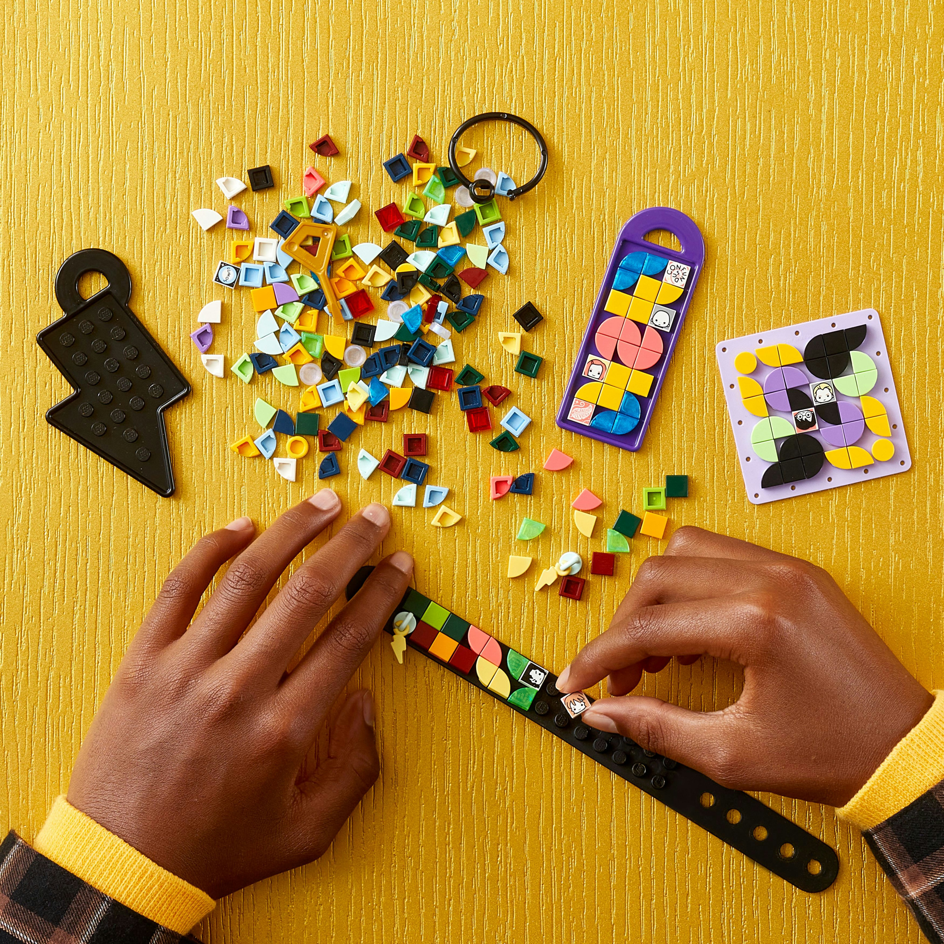Ręce składające bransoletkę z kolorowych klocków Lego na żółtym tle. Obecne są inne elementy i czarny kształt.