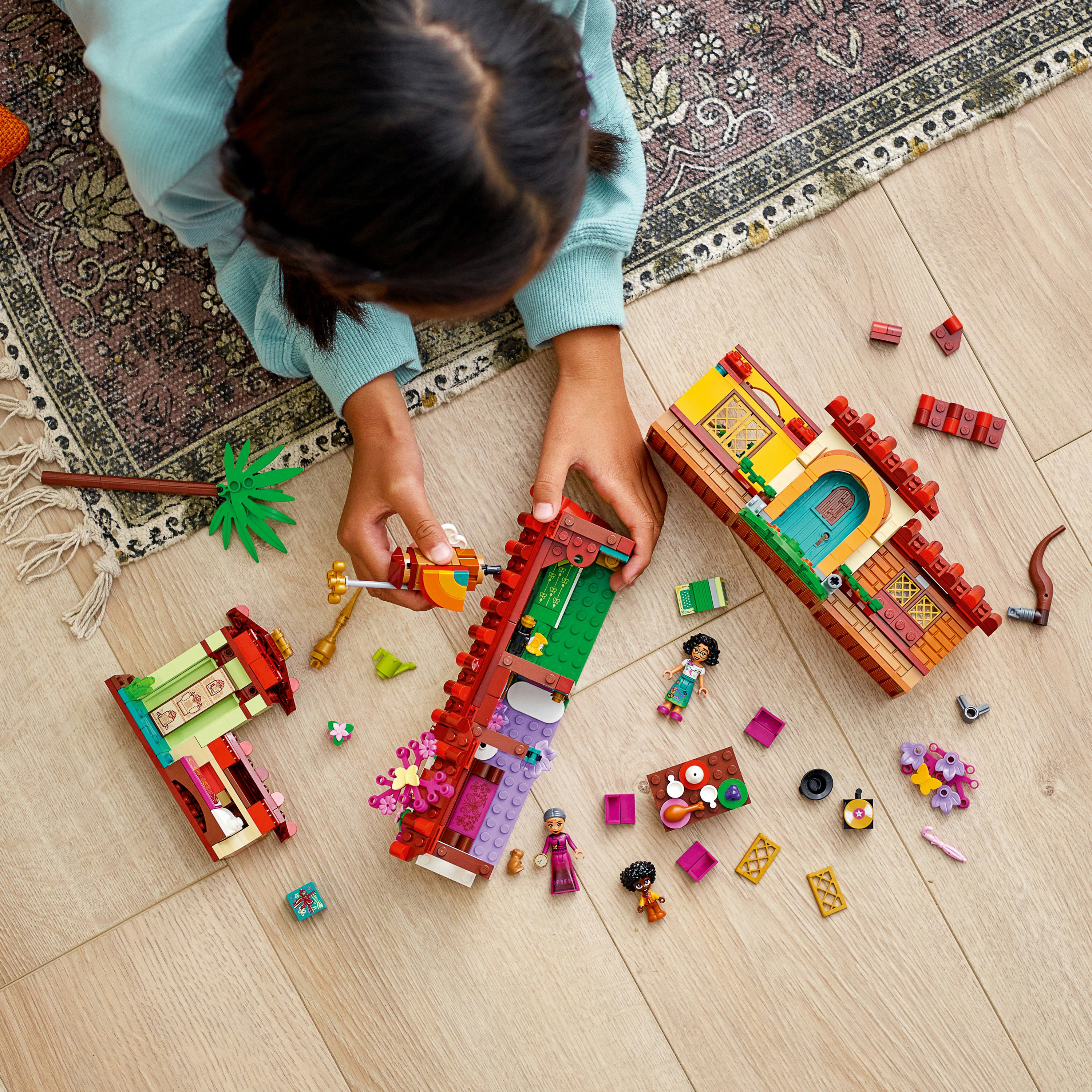 Ein Kind baut ein Lego-Set mit einem farbenfrohen Haus mit mehreren Abschnitten und Lego-Figuren auf einem Holzboden.