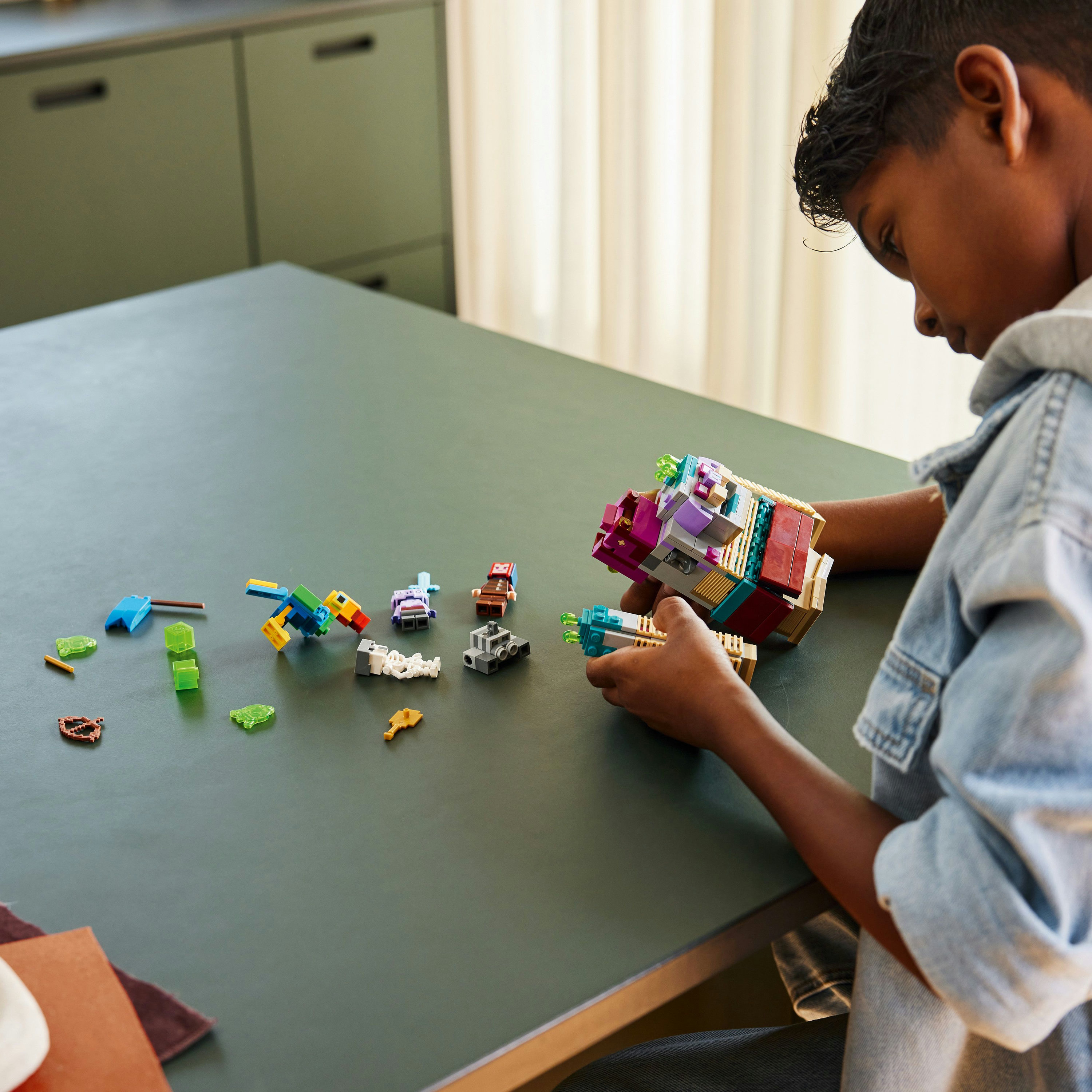 Ein Kind, das Lego-Teile auf einem dunkelgrünen Tisch zusammensetzt, mit anderen Lego-Elementen verstreut.