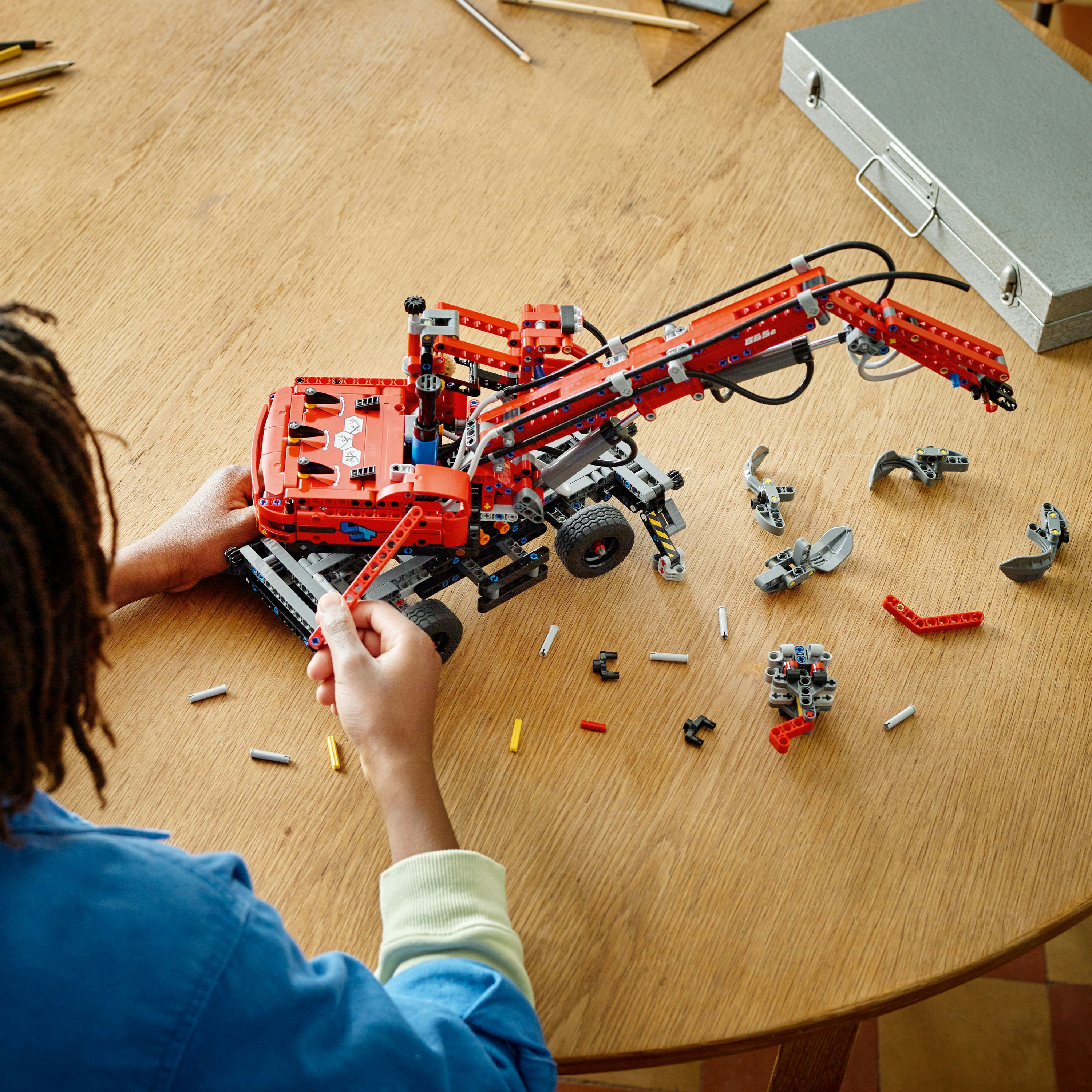 Eine Person montiert einen rot-grauen LEGO Technic Kran auf einem Holztisch. Teile sind verstreut.