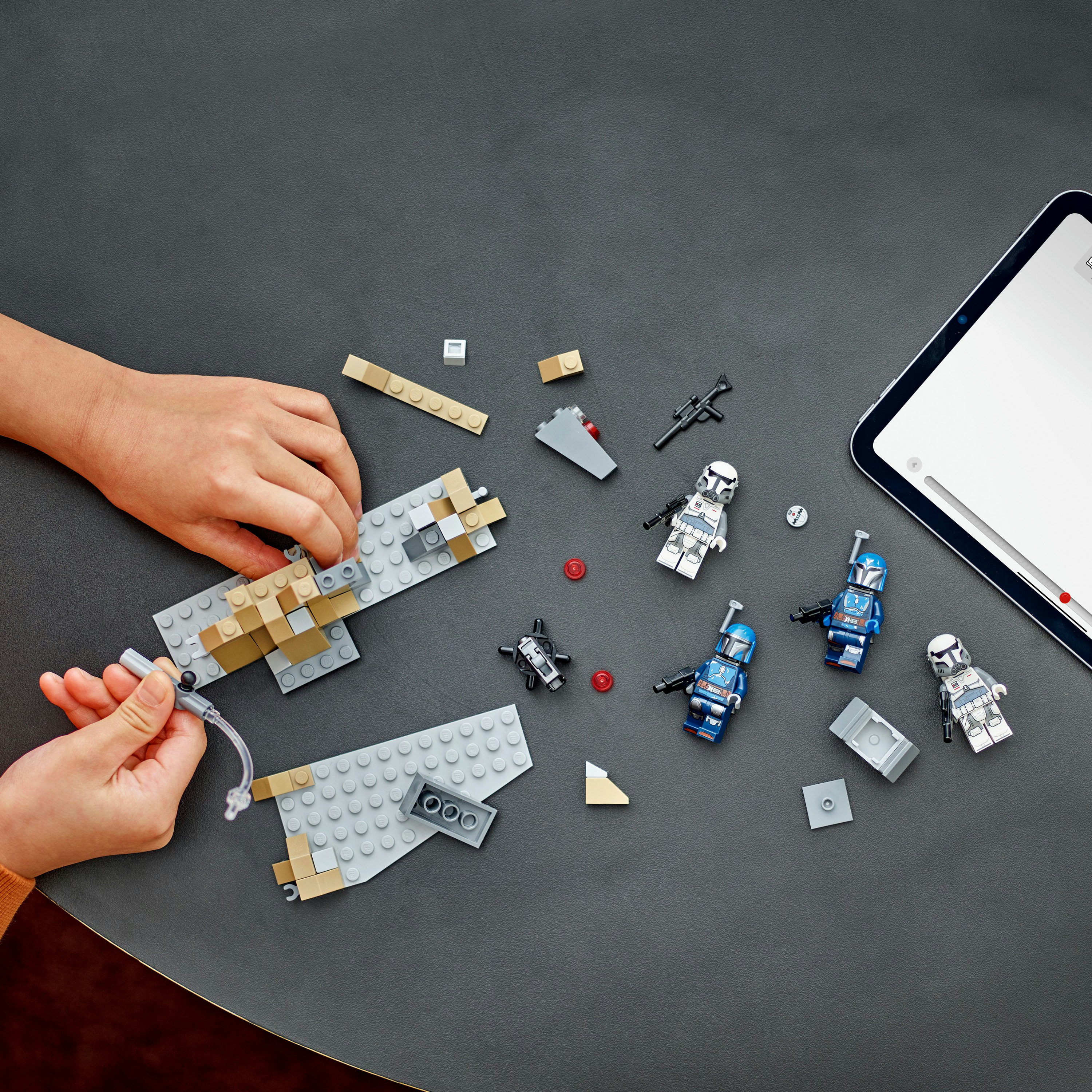 Ręce składające klocki Lego na ciemnej powierzchni, z figurkami Lego w pobliżu i tabletem.