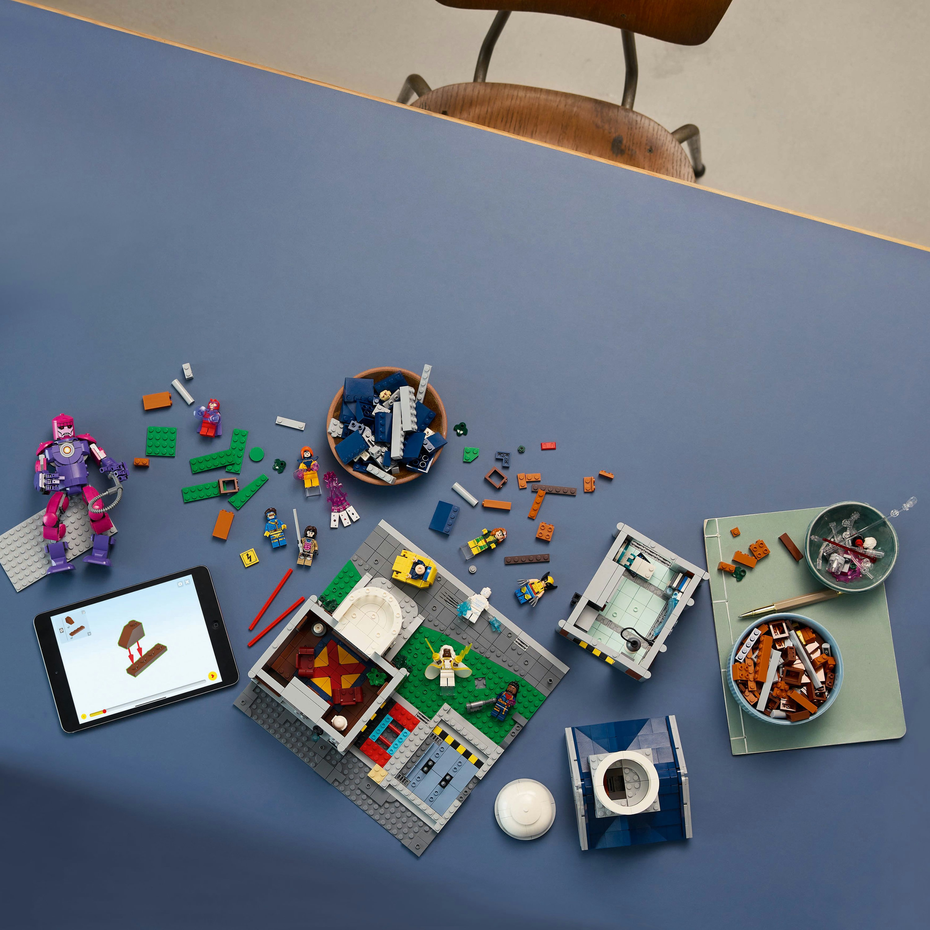 Lego parçaları, modelleri ve figürlerle kaplı bir masa. Talimatların yer aldığı bir tablet görülebilir.
