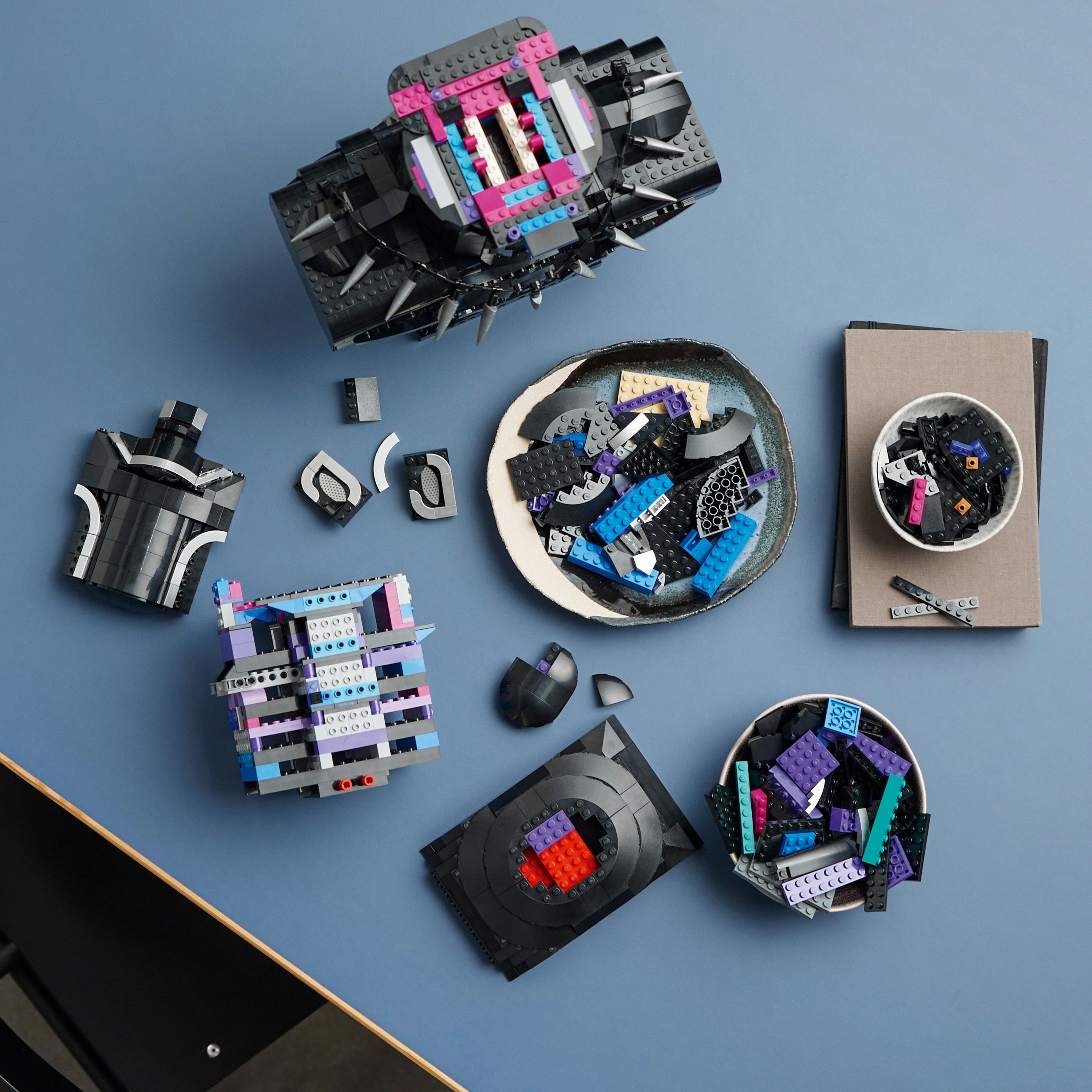 Elementy zestawu LEGO są ułożone na niebieskiej powierzchni. Zawiera czarne części, płytkę z kolorowymi detalami i inne obiekty.