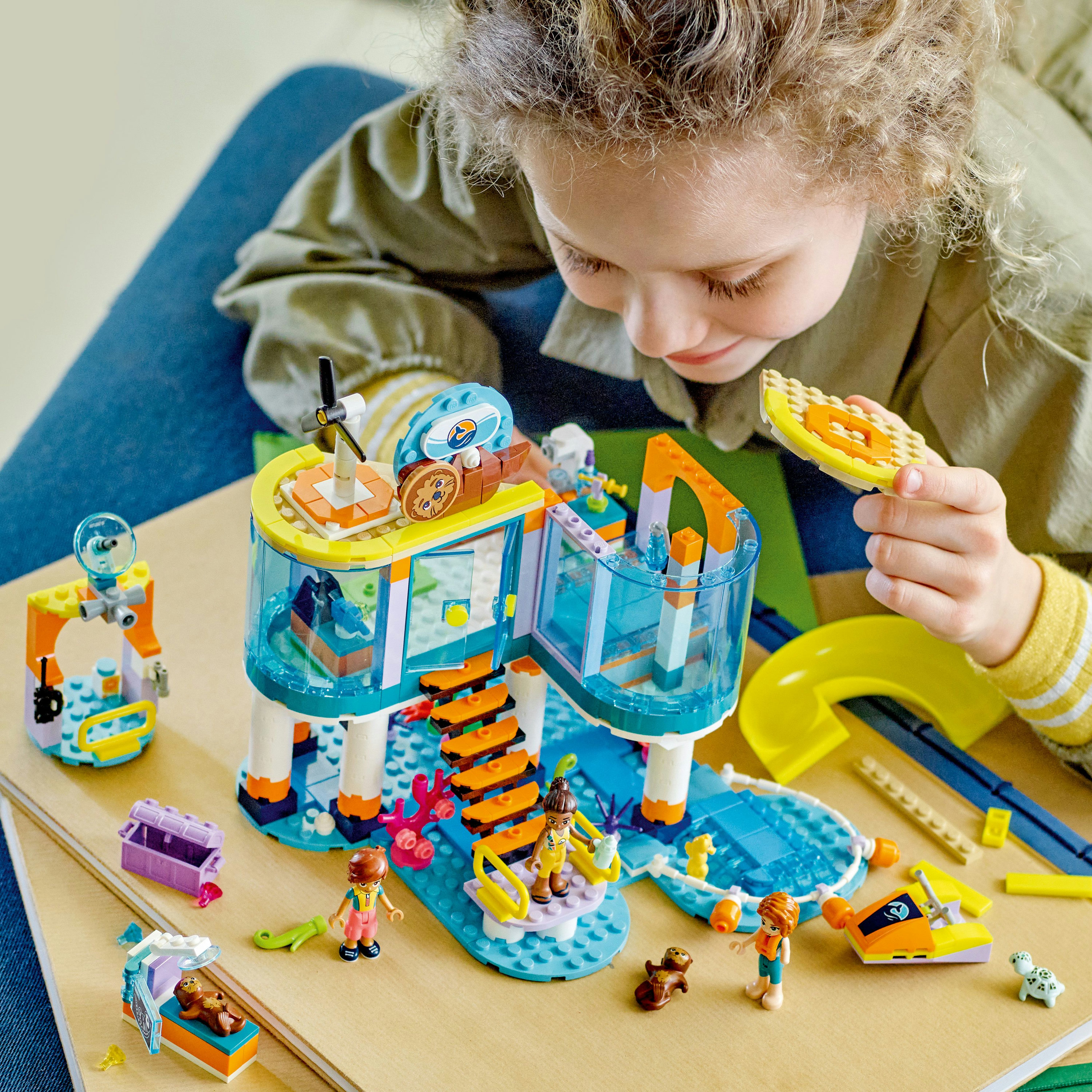 Ein Mädchen spielt mit einem Lego-Set, das einen Pool, Rutschen und Figuren zeigt. Das Set hat ein farbenfrohes Design.