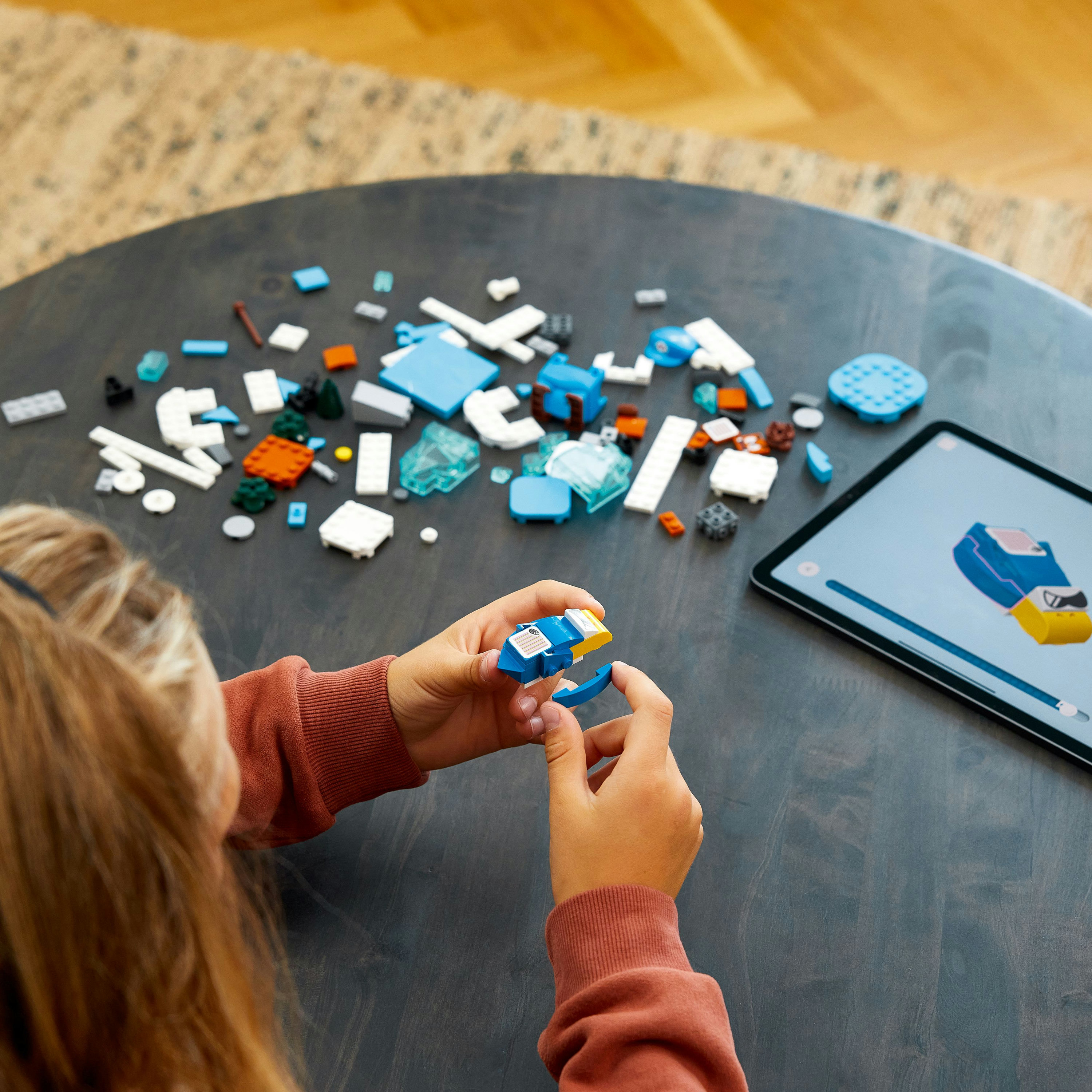 Eine Person baut ein blaues und gelbes LEGO-Modell auf einem dunklen Holztisch. Es gibt verstreute LEGO-Teile und ein Tablet.
