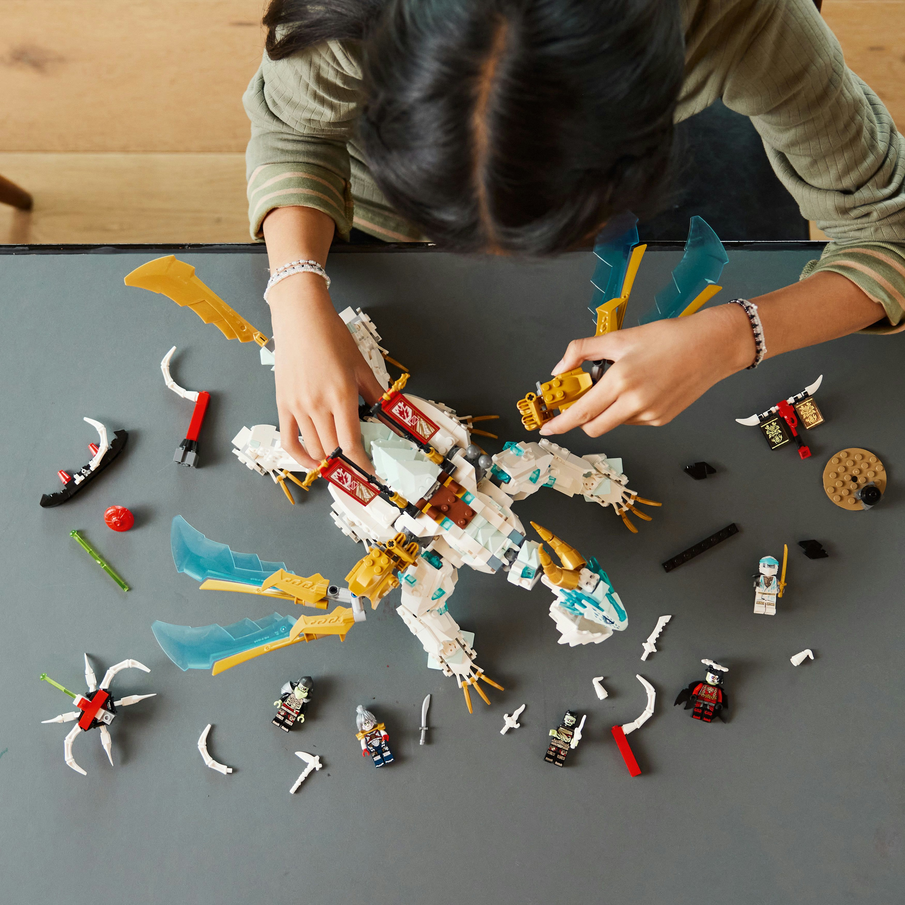 Eine Person baut einen weißen und goldenen LEGO-Drachen auf einer grauen Oberfläche zusammen, umgeben von Elementen und Minifiguren.