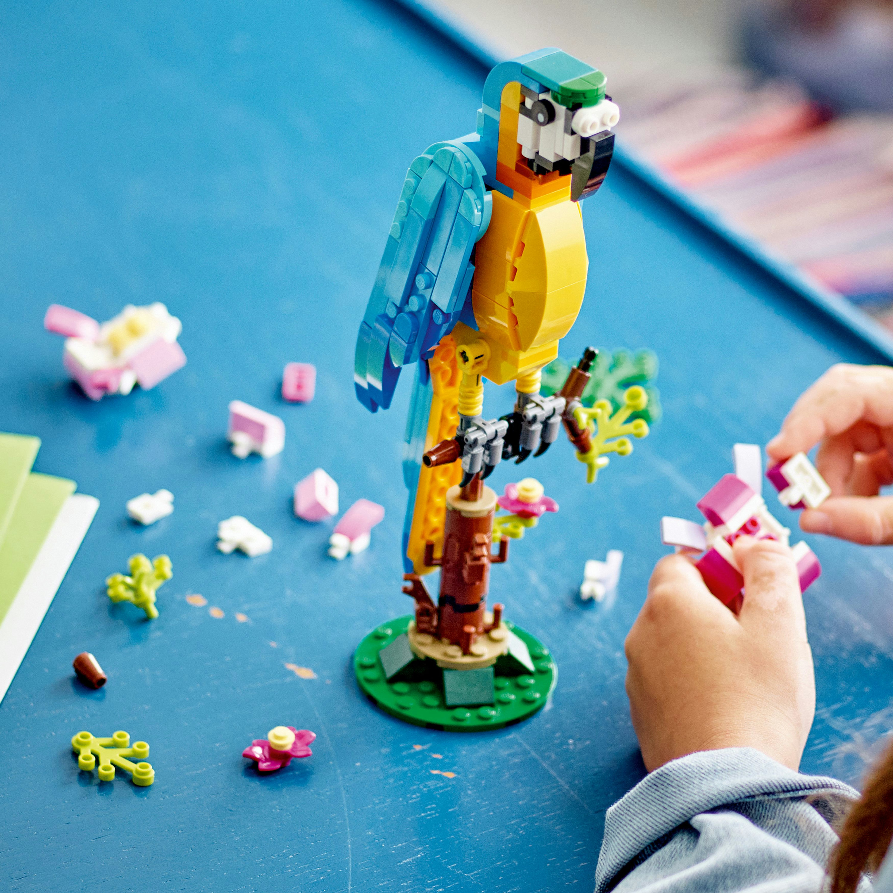 Osoba buduje papugę Lego. Papuga jest niebieska, żółta i zielona, siedzi na brązowym pniu drzewa.