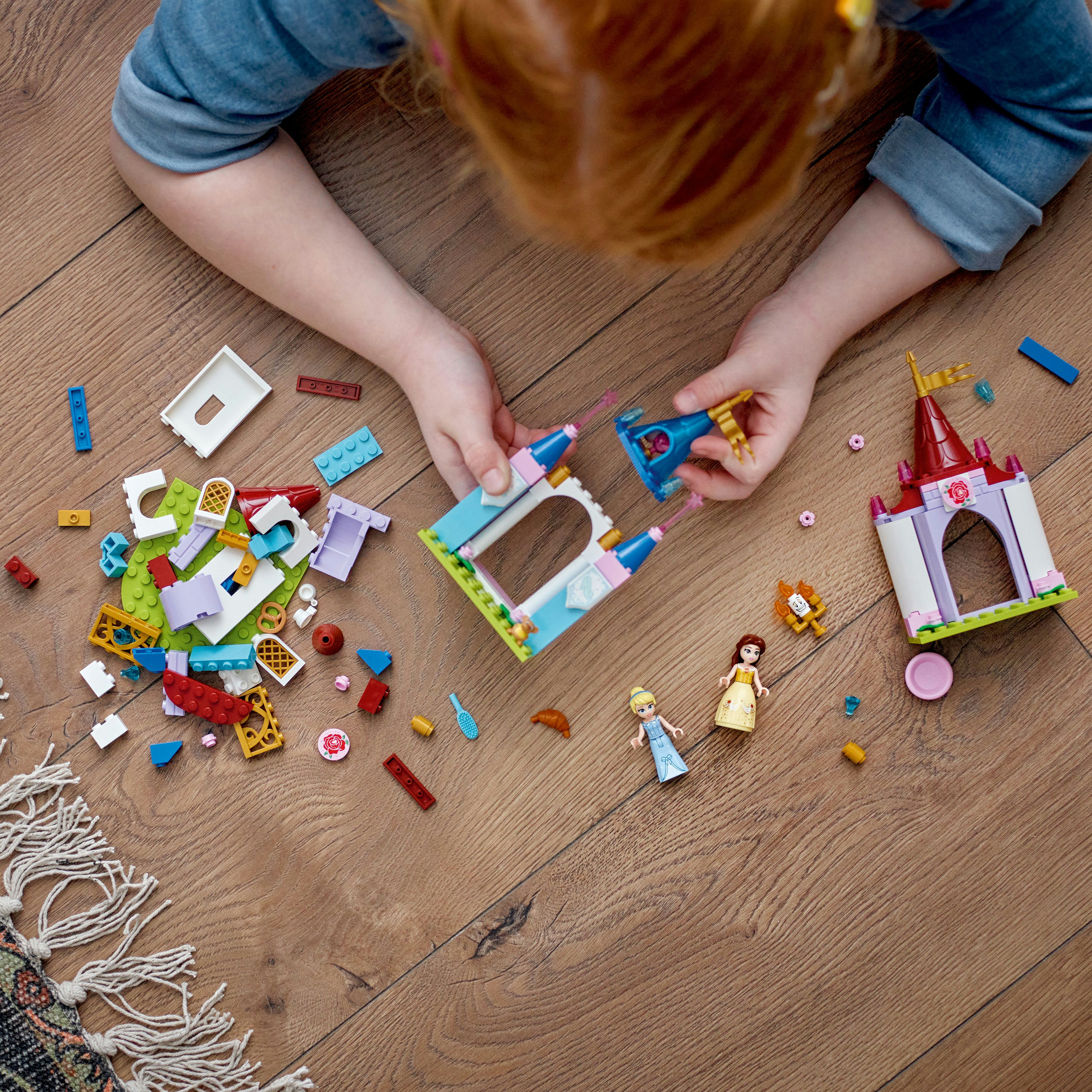 Ein Kind spielt mit Lego-Teilen. Auf einem Holzboden liegen eine Burg und andere Steine verstreut. Ein Mädchen hält ein Teil.