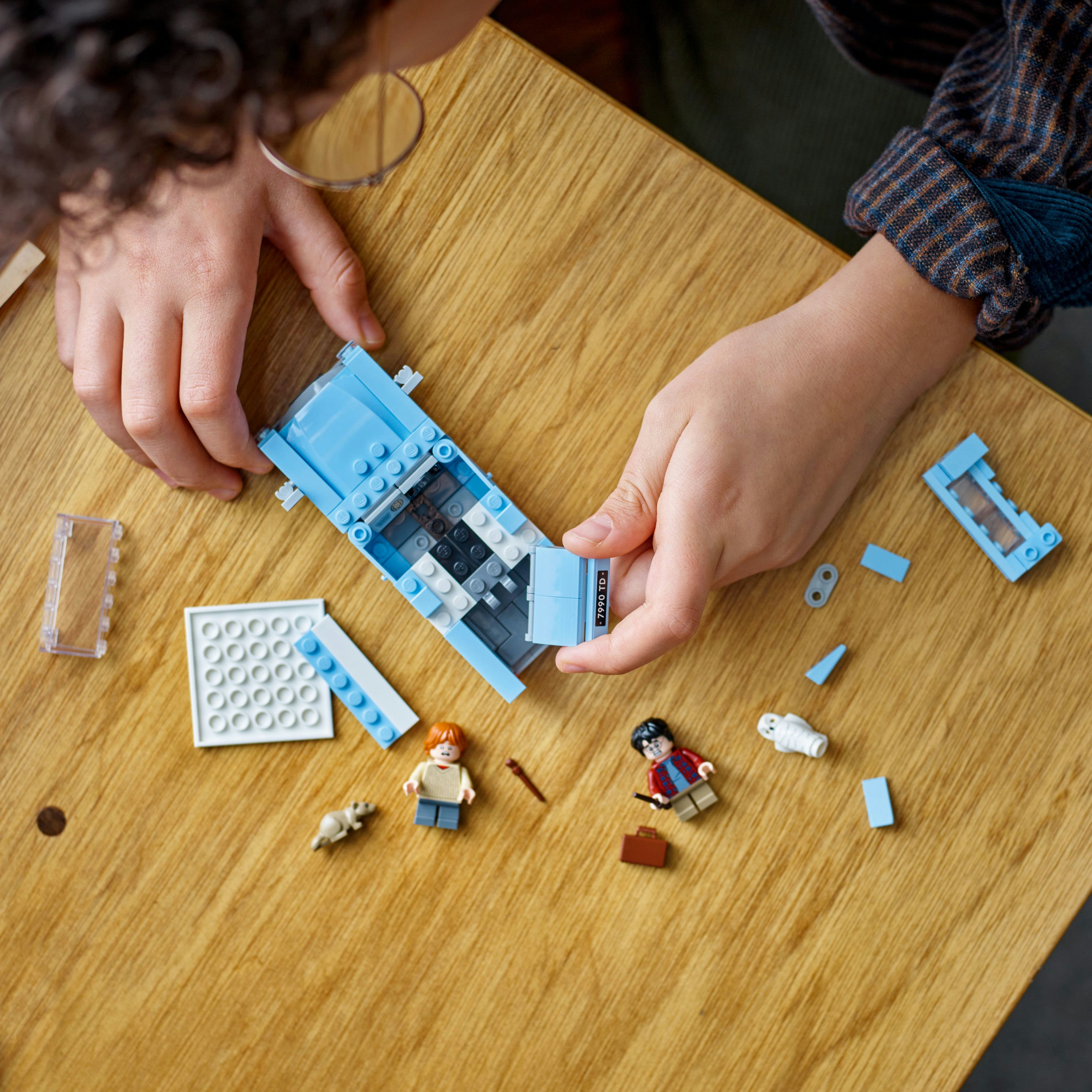 Osoba składa niebieski samochód Lego. Kilka klocków Lego i figurek jest rozrzuconych na stole.