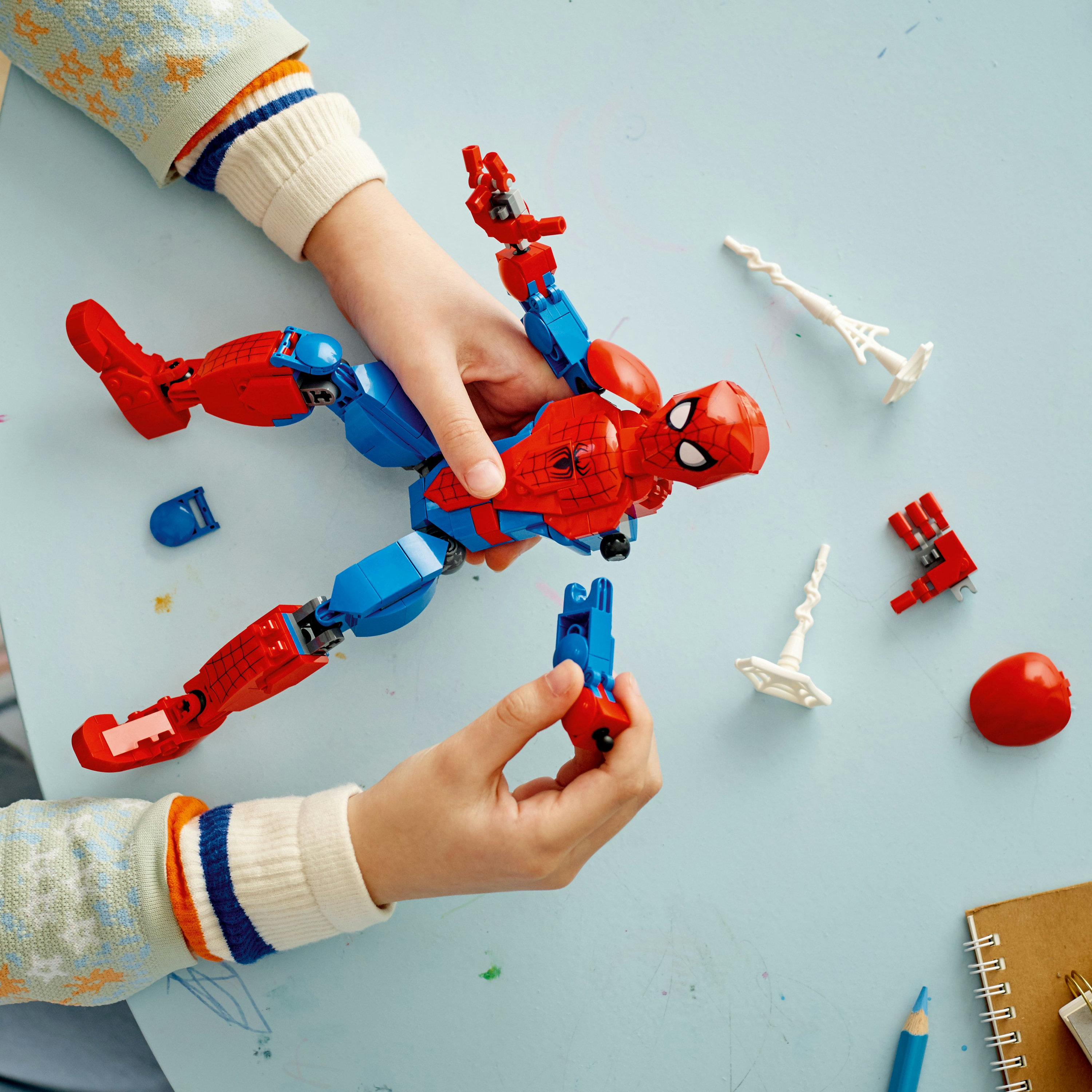 Hände, die eine Spiderman-Lego-Figur auf einer hellblauen Oberfläche zusammensetzen. Verschiedene Teile sind verstreut.