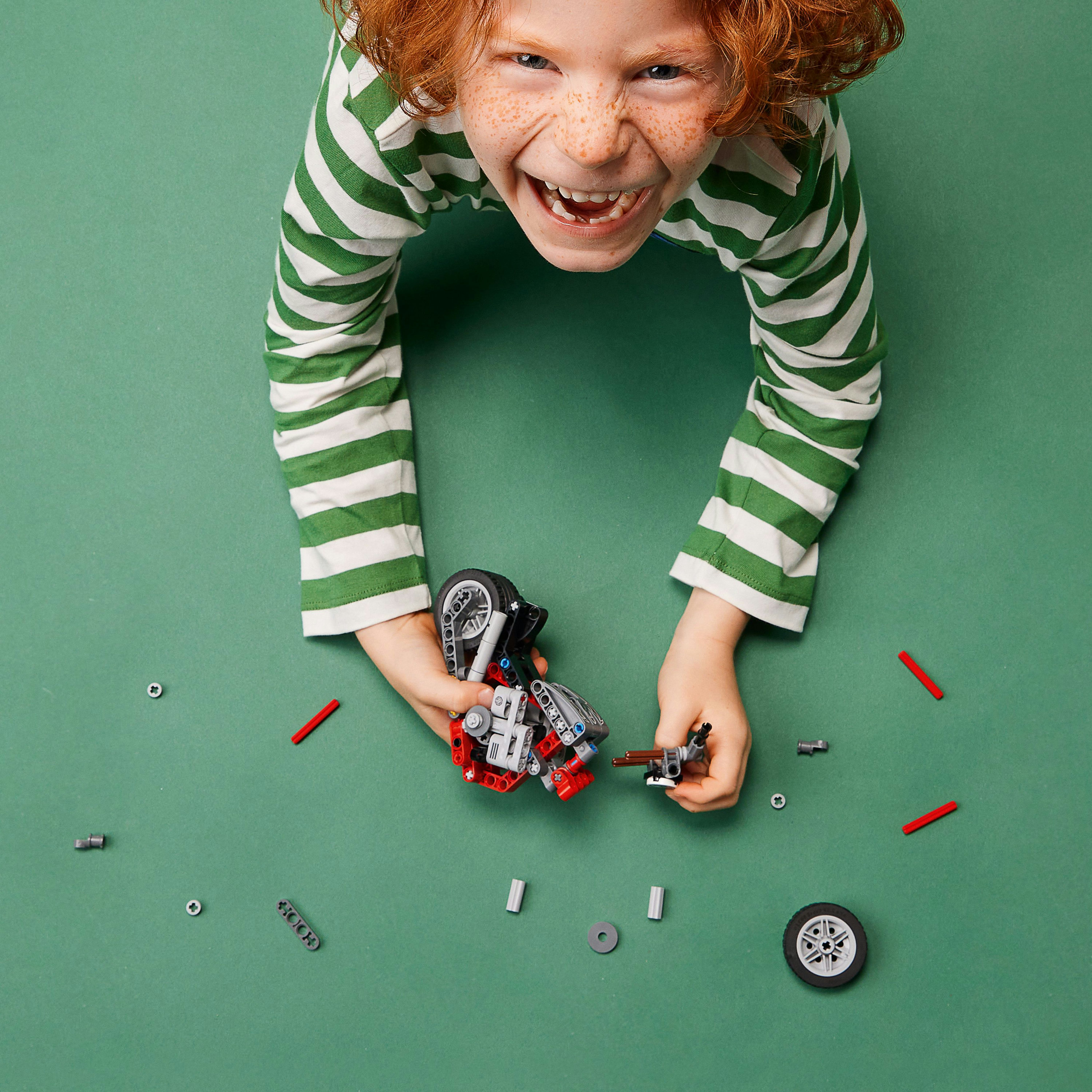 Kızıl saçlı bir çocuk, yeşil bir yüzeyde bir Lego motosiklet toplarken gülümsüyor.