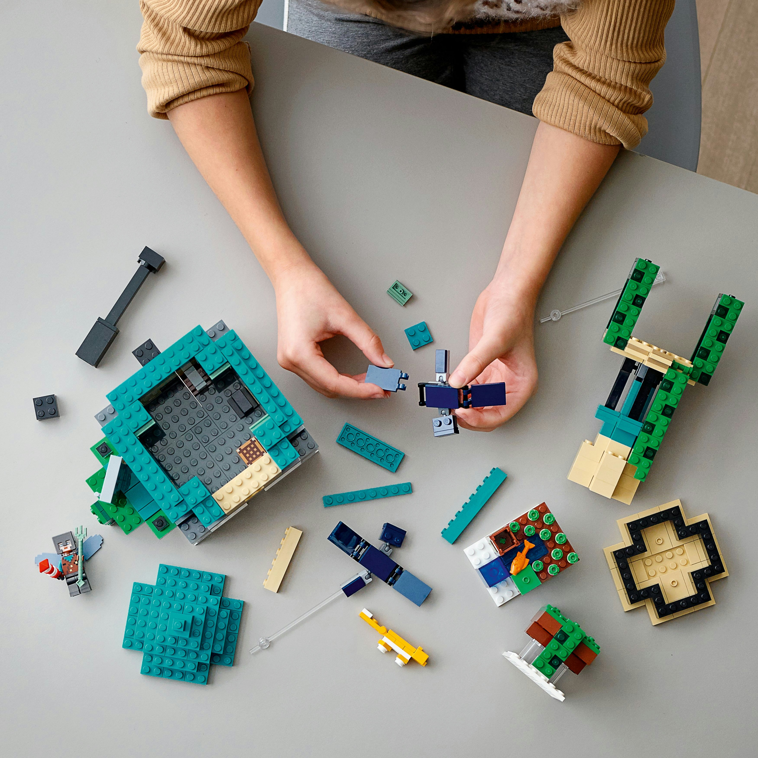 Mani che assemblano una scena LEGO Minecraft con figure e mattoncini su un tavolo.