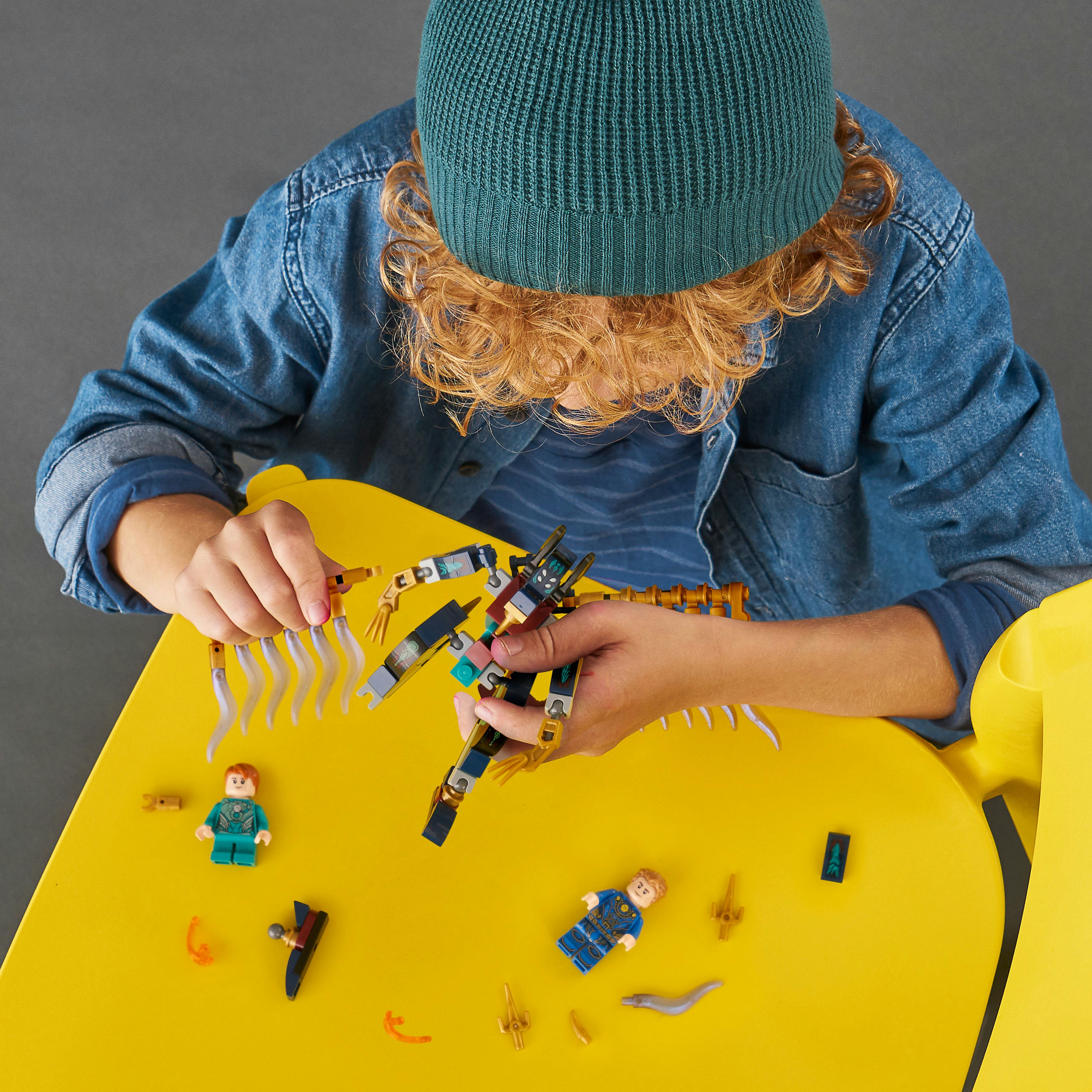 Ein Kind mit grüner Mütze baut ein Lego-Set auf einem gelben Tisch zusammen. Lego-Figuren und Teile liegen herum.