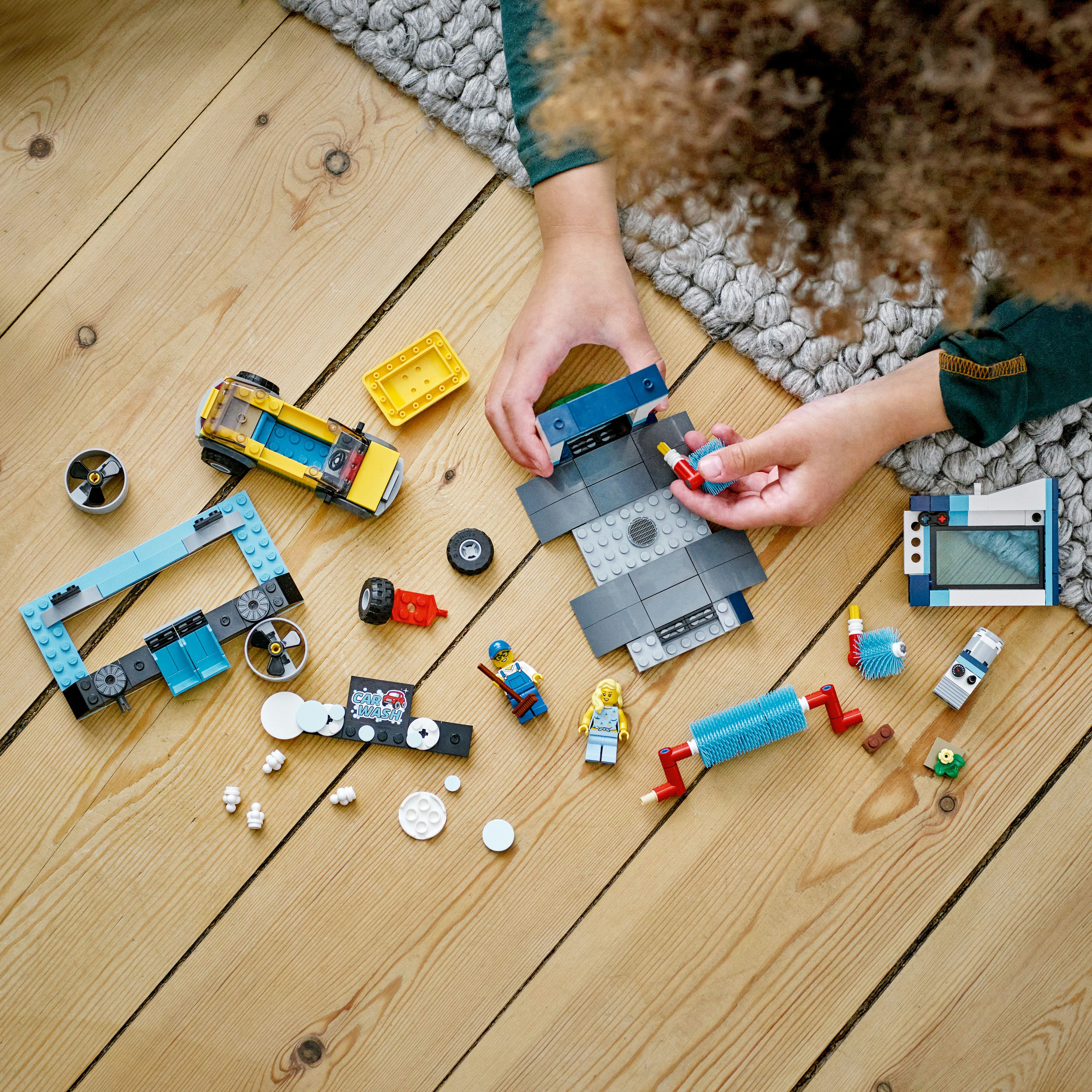 Die Hände eines Kindes setzen LEGO-Teile auf einem Holzboden zusammen. Verschiedene LEGO-Komponenten sind verstreut.